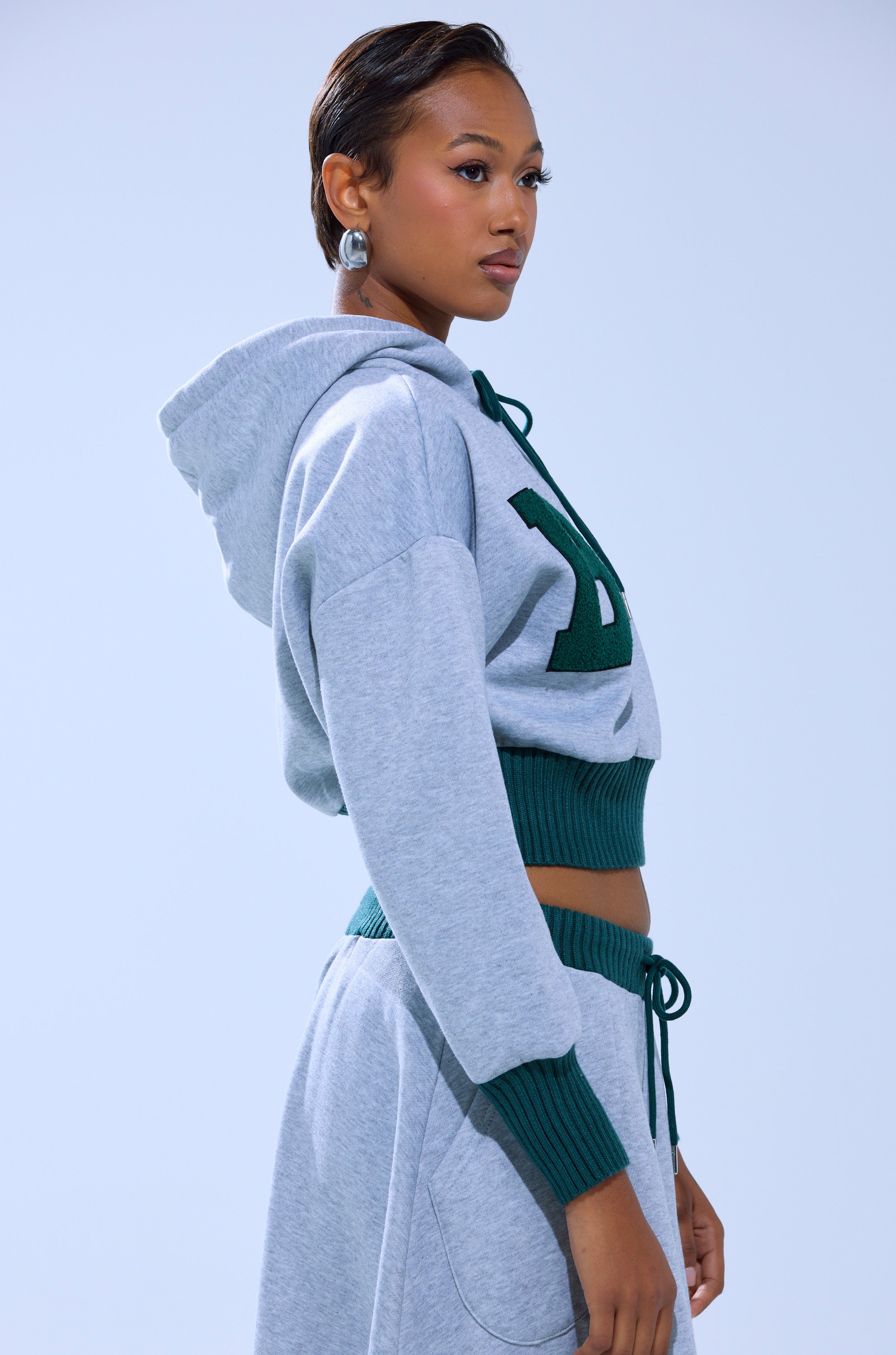 A woman in profile wears the COLLEGIATE FRENCH TERRY HOODED SWEATSHIRT in light gray with green trim and matching sweatpants. Her hair is slicked back, she has statement earrings, and the background is plain light blue.
