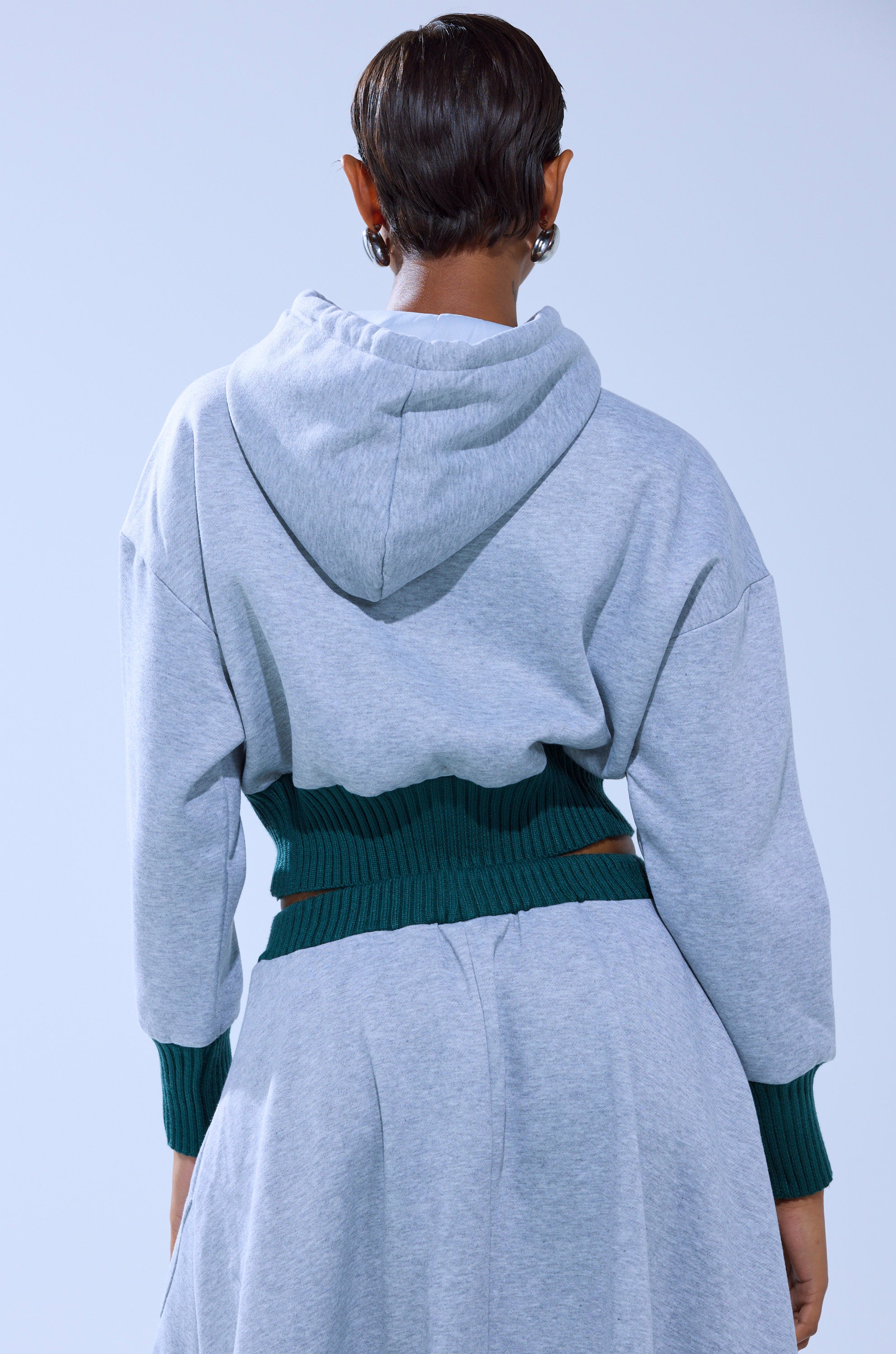 A person with short hair is seen from behind in a light gray COLLEGIATE FRENCH TERRY HOODED SWEATSHIRT featuring green ribbed waist and cuffs, paired with a matching skirt. The background is plain light blue.