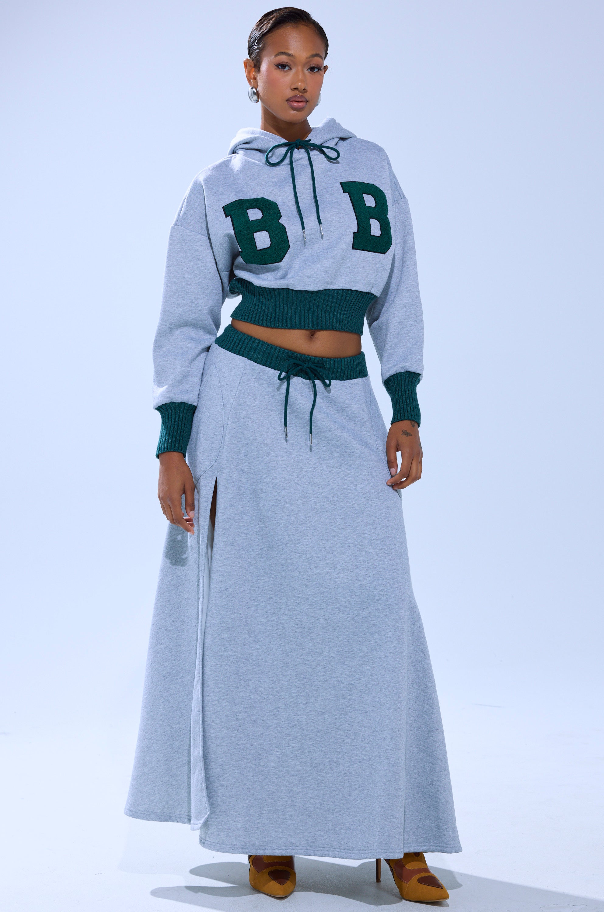 A woman wears the COLLEGIATE FRENCH TERRY HOODED SWEATSHIRT in light gray with green "BB" letters, paired with a matching drawstring maxi skirt and brown heels, all against a plain light background.