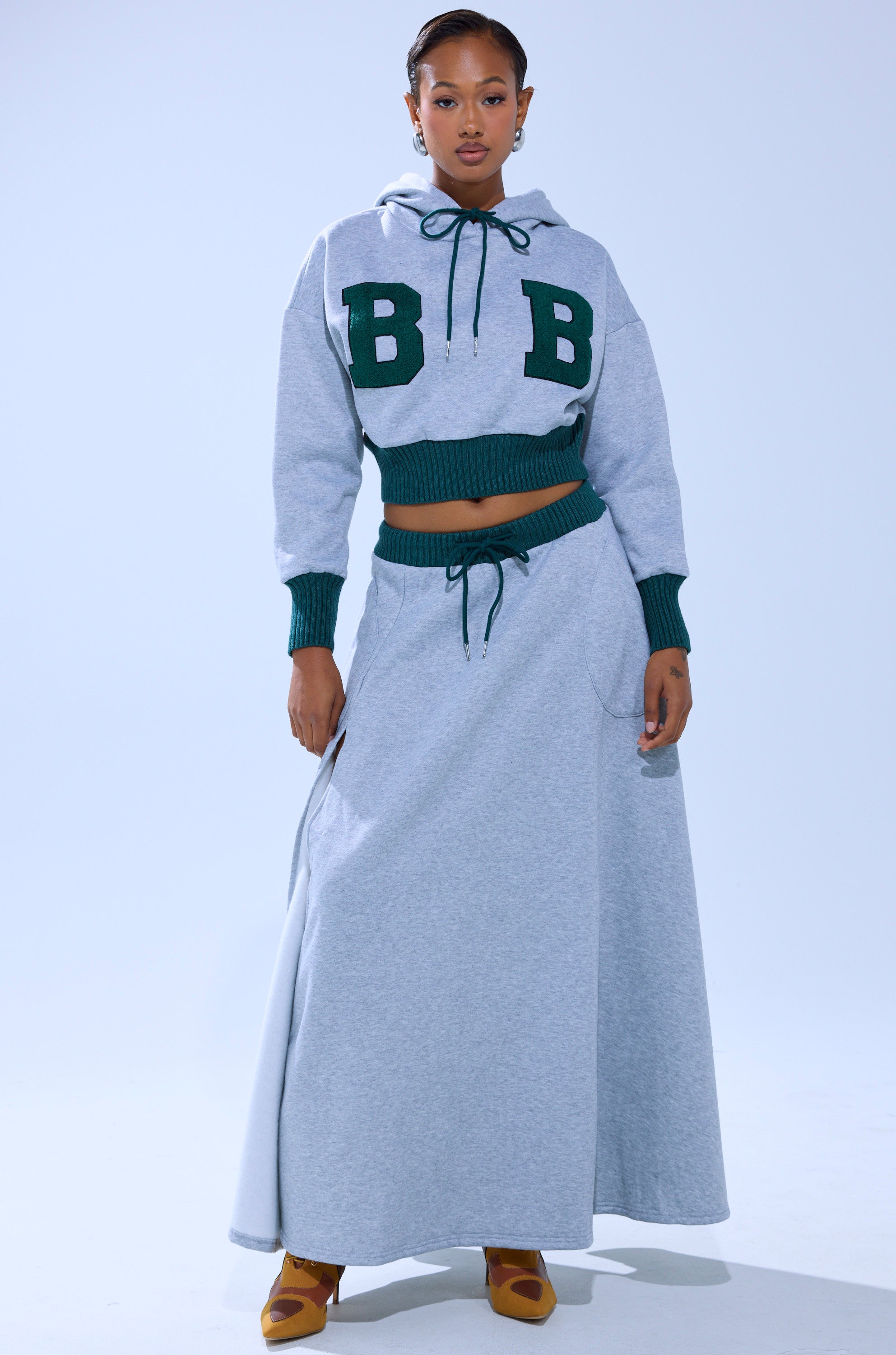 A woman wears the COLLEGIATE FRENCH TERRY HOODED SWEATSHIRT—gray with green cuffs and bold "B B" letters—paired with a matching long gray skirt and tan heels against a light background.