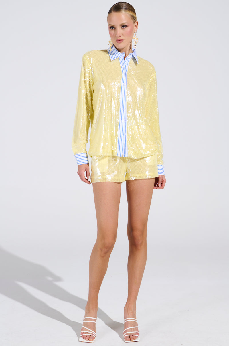 A woman poses in a studio wearing the DOLLY SEQUIN SHORT—a shiny yellow, high-rise short with an elastic waistband—styled with a matching long-sleeve shirt with blue and white striped collar and cuffs, white strappy heels, and large hoop earrings.