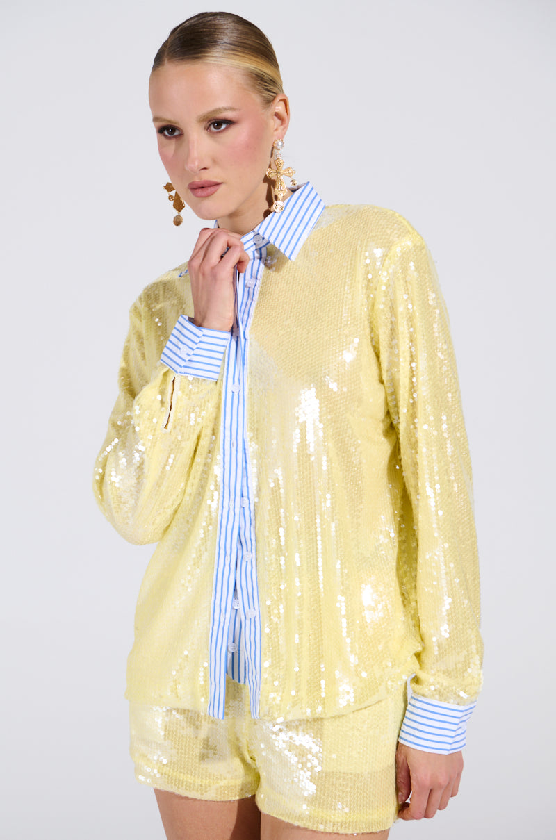 A woman in the DOLLY SEQUIN BUTTON DOWN TOP, with a shimmering sequin overlay and blue and white striped cuffs and collar, poses confidently against a plain light background. She has slicked-back hair and statement earrings. 0