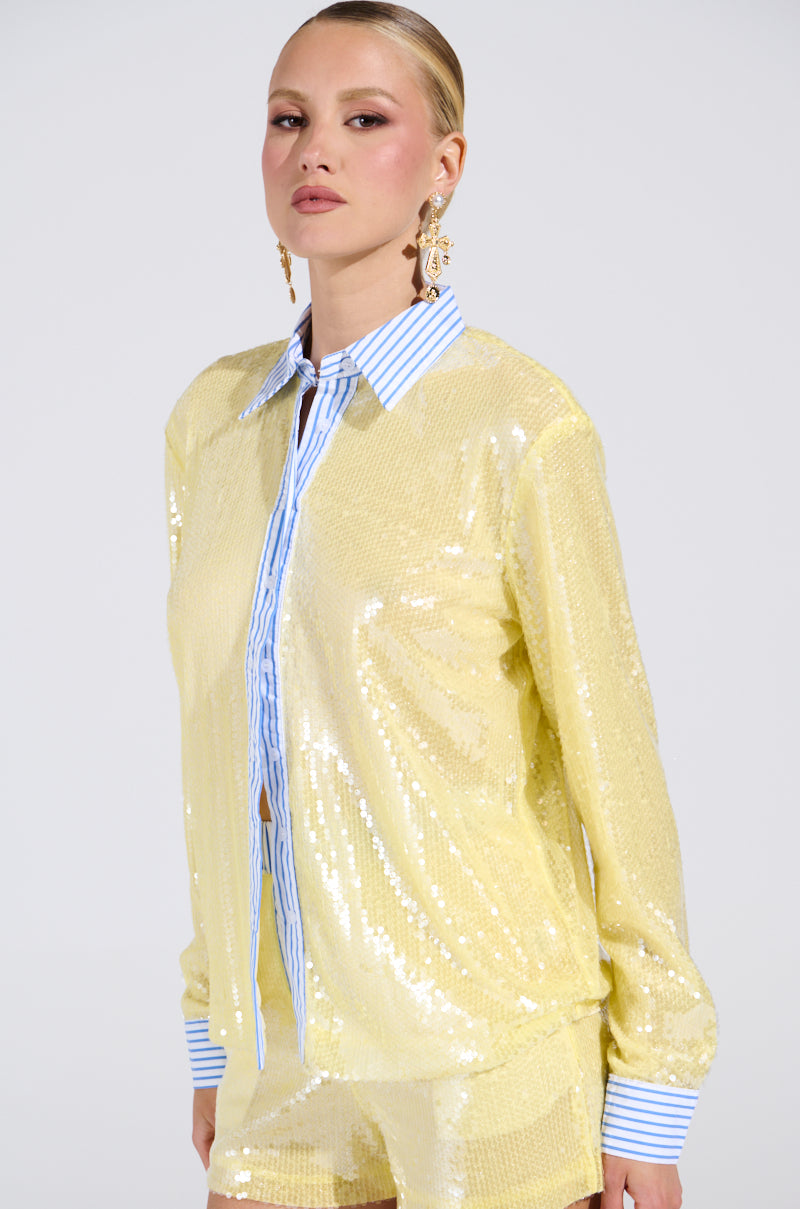 A woman wears the shiny yellow DOLLY SEQUIN BUTTON DOWN TOP over a blue and white striped collared shirt, paired with matching yellow shorts. She styles her hair slicked back and accessorizes with gold statement earrings. 1