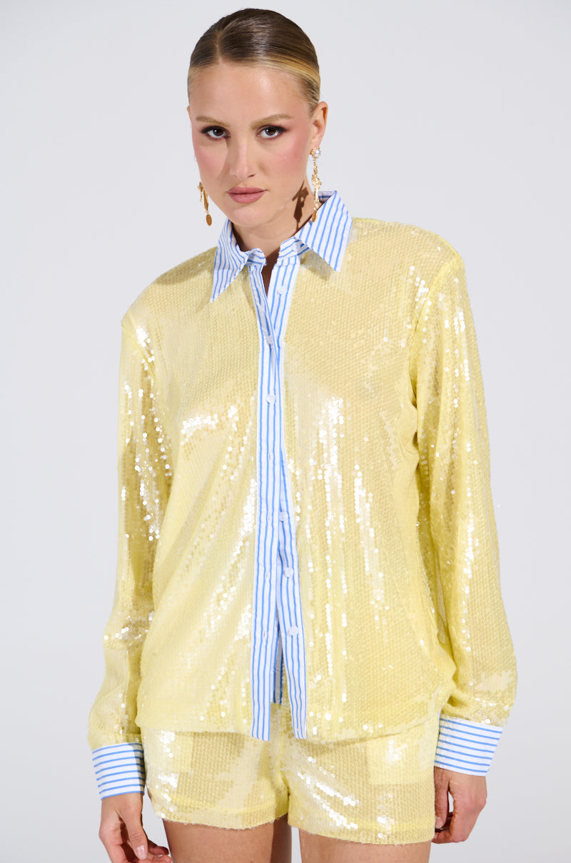 A woman with slicked-back hair wears the DOLLY SEQUIN BUTTON DOWN TOP—a yellow sequin overlay shirt with blue and white striped collar and cuffs—paired with matching shorts, standing against a plain light background. 2