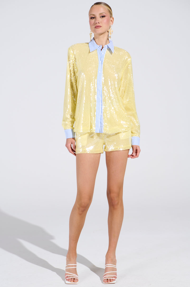 A woman wears the DOLLY SEQUIN BUTTON DOWN TOP—a loose, shiny yellow sequin shirt with blue-and-white striped cuffs and collar—with matching sequin shorts, white strappy heels, and large hoop earrings against a plain background. 5