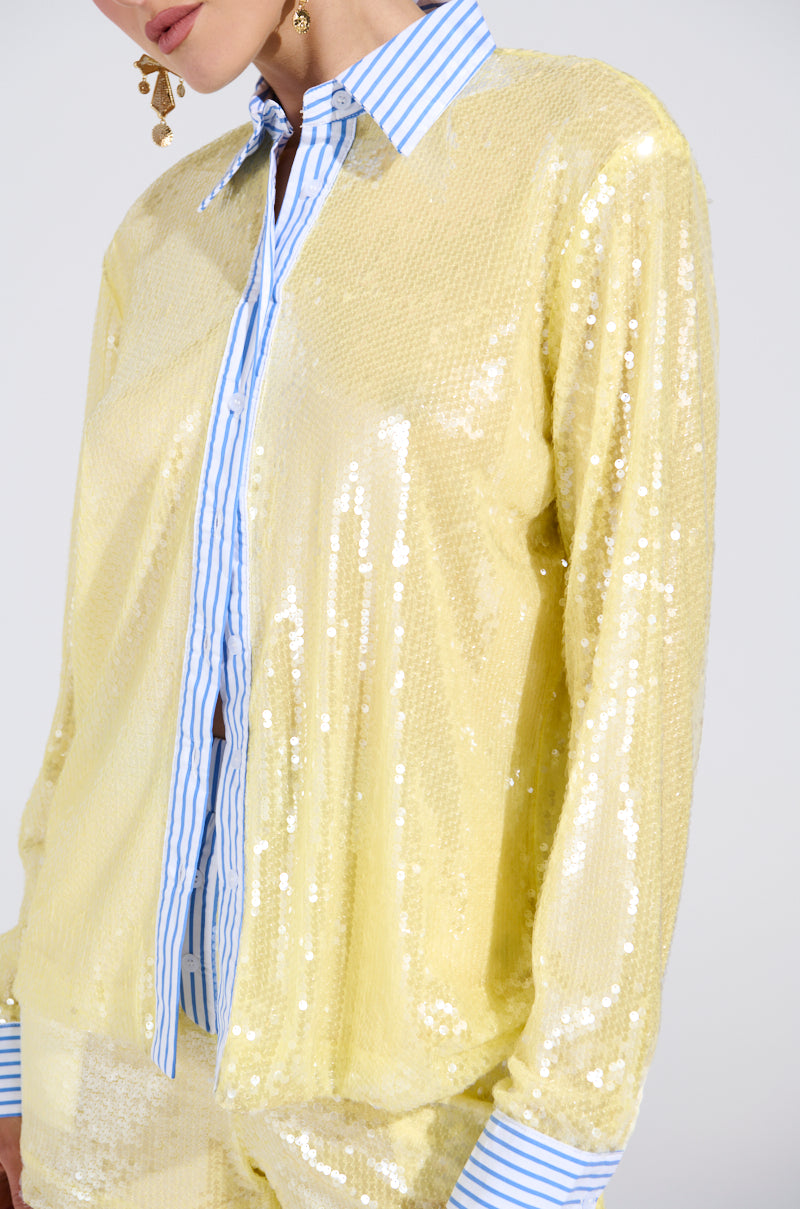 A person wears the DOLLY SEQUIN BUTTON DOWN TOP, a shiny yellow sequin long-sleeve shirt layered over a blue and white striped collared shirt with matching cuffs. Gold earrings complete the look, while their face is partially out of frame. 7