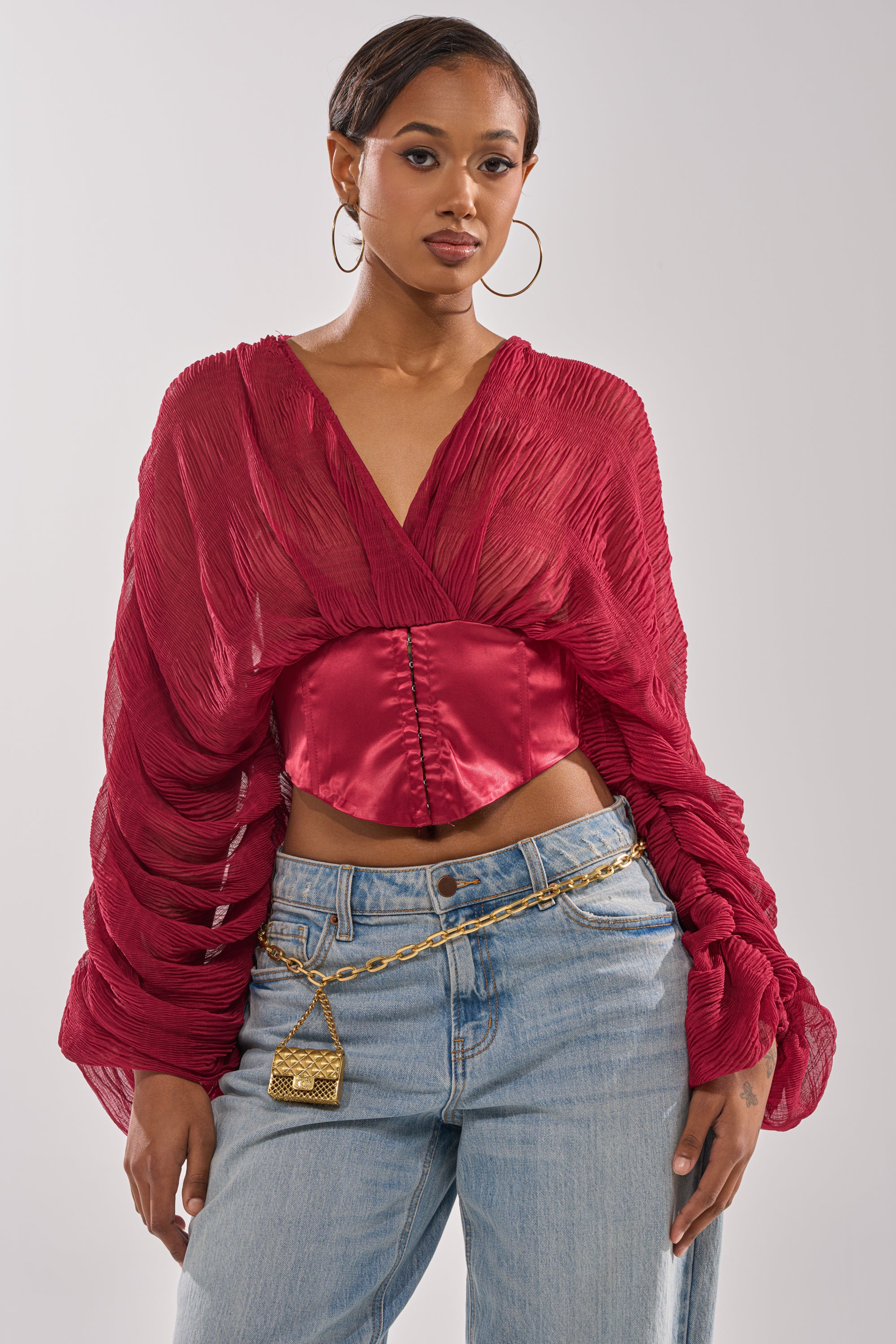 A woman poses against a plain background wearing the GIRLY PLZ BLOUSE—red, sheer chiffon with ruched balloon sleeves and a shiny corset-style bodice—paired with light blue jeans, large hoop earrings, and a gold chain belt with a lock pendant. 0
