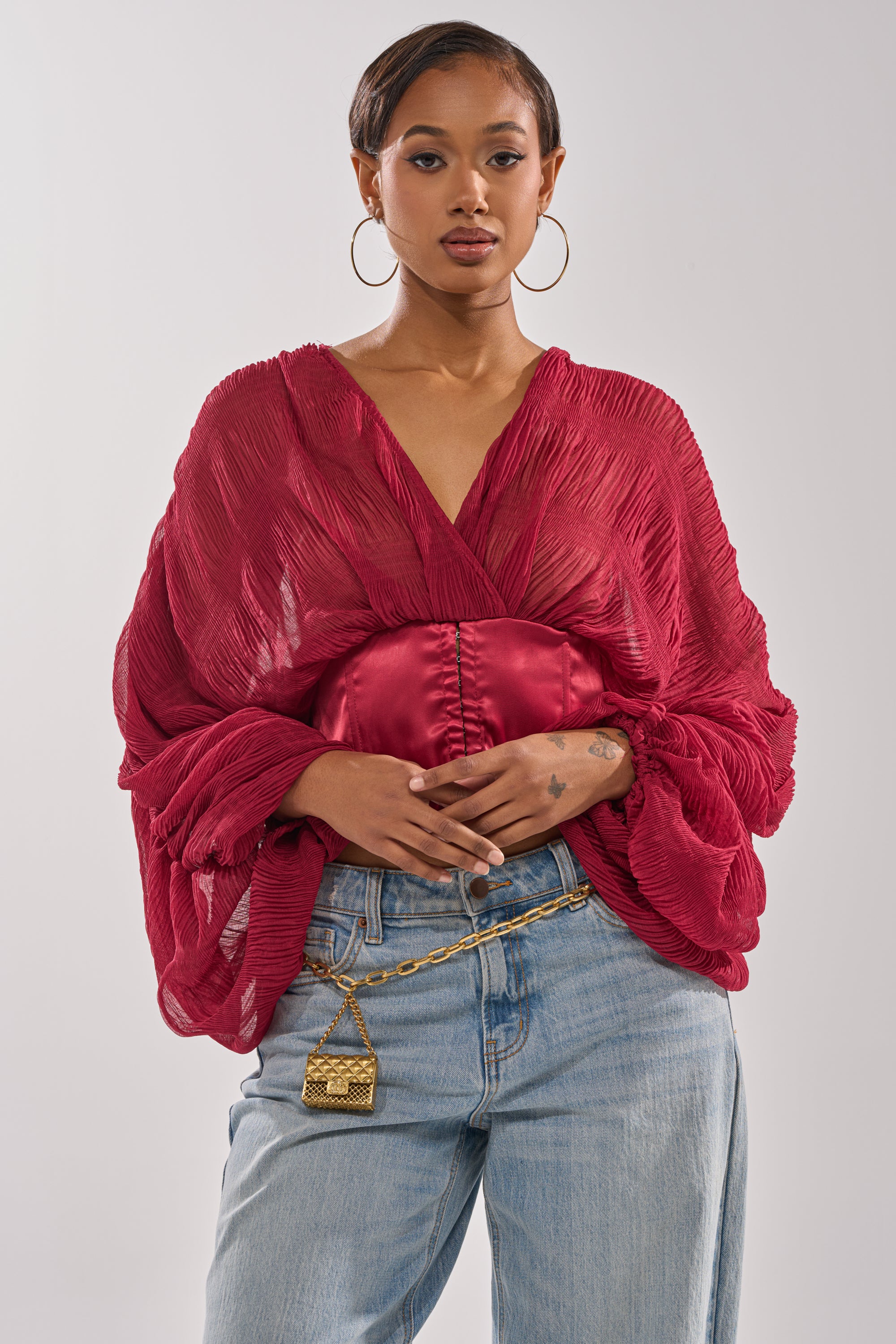 A woman wears the GIRLY PLZ BLOUSE—a sheer chiffon top with ruched balloon sleeves—paired with light blue jeans, large hoop earrings, and a gold chain belt with a small handbag, standing against a plain background. 1