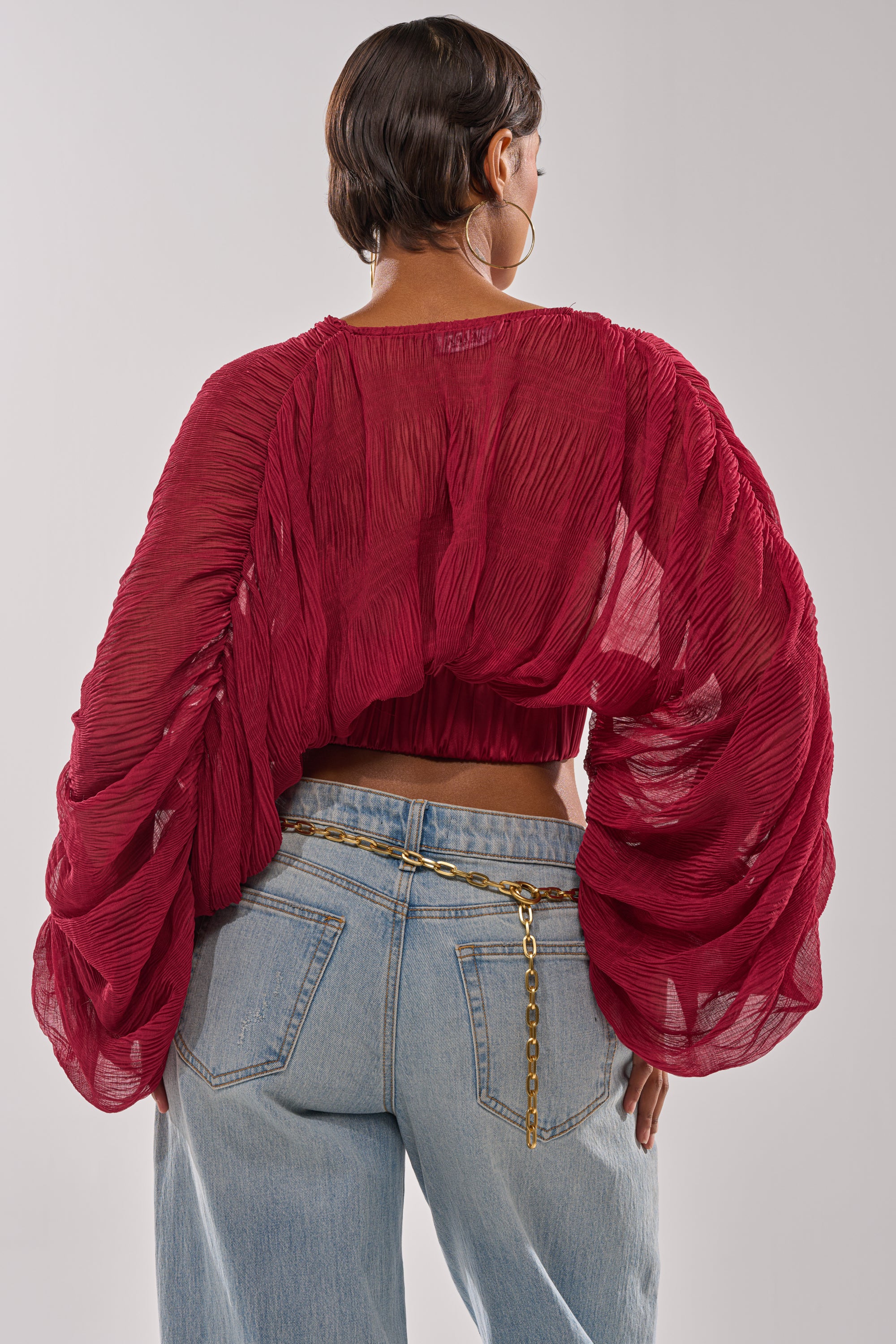 A person with short dark hair, back to the camera, wears the GIRLY PLZ BLOUSE—a sheer chiffon top with billowy sleeves—paired with light blue jeans and a gold chain belt, set against a neutral background. 3