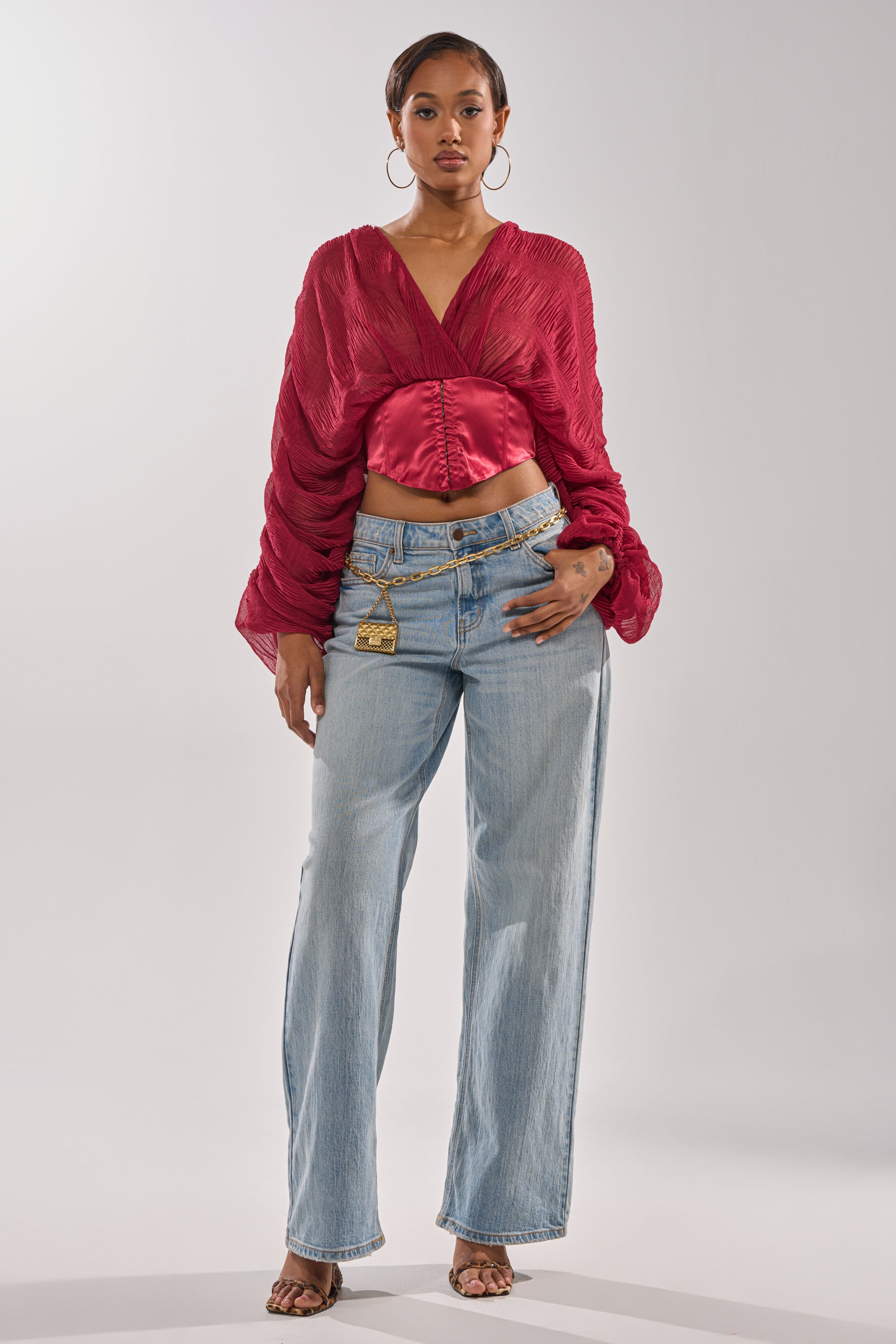A woman with short hair and a neutral expression faces forward, wearing the GIRLY PLZ BLOUSE in deep red with ruched balloon sleeves, paired with light blue high-waisted jeans, hoop earrings, and heeled sandals. 4