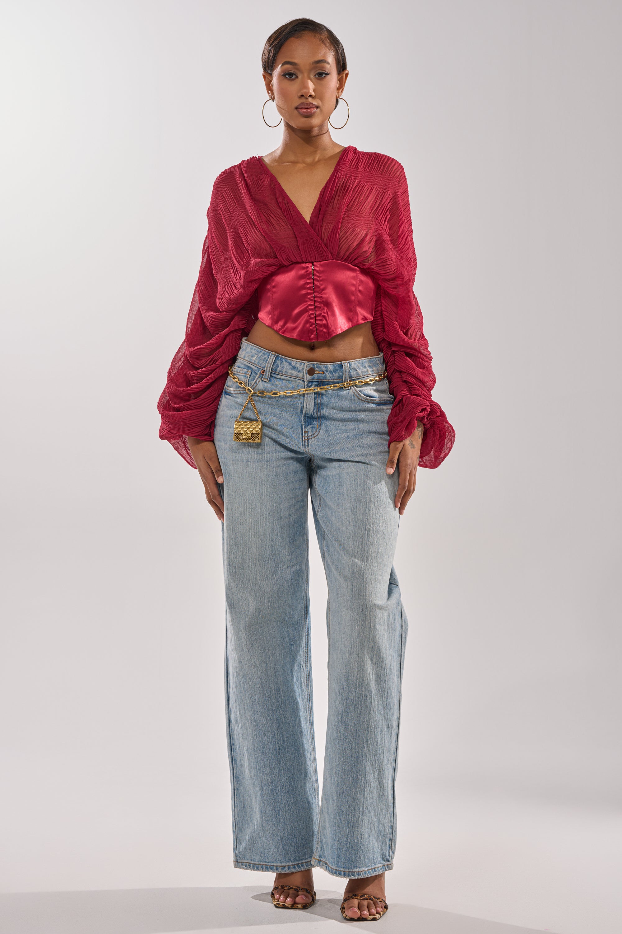A woman stands confidently against a plain background wearing the GIRLY PLZ BLOUSE in red chiffon with ruched balloon sleeves, paired with light blue wide-leg jeans, gold hoop earrings, layered gold belts, and open-toed heels. 5