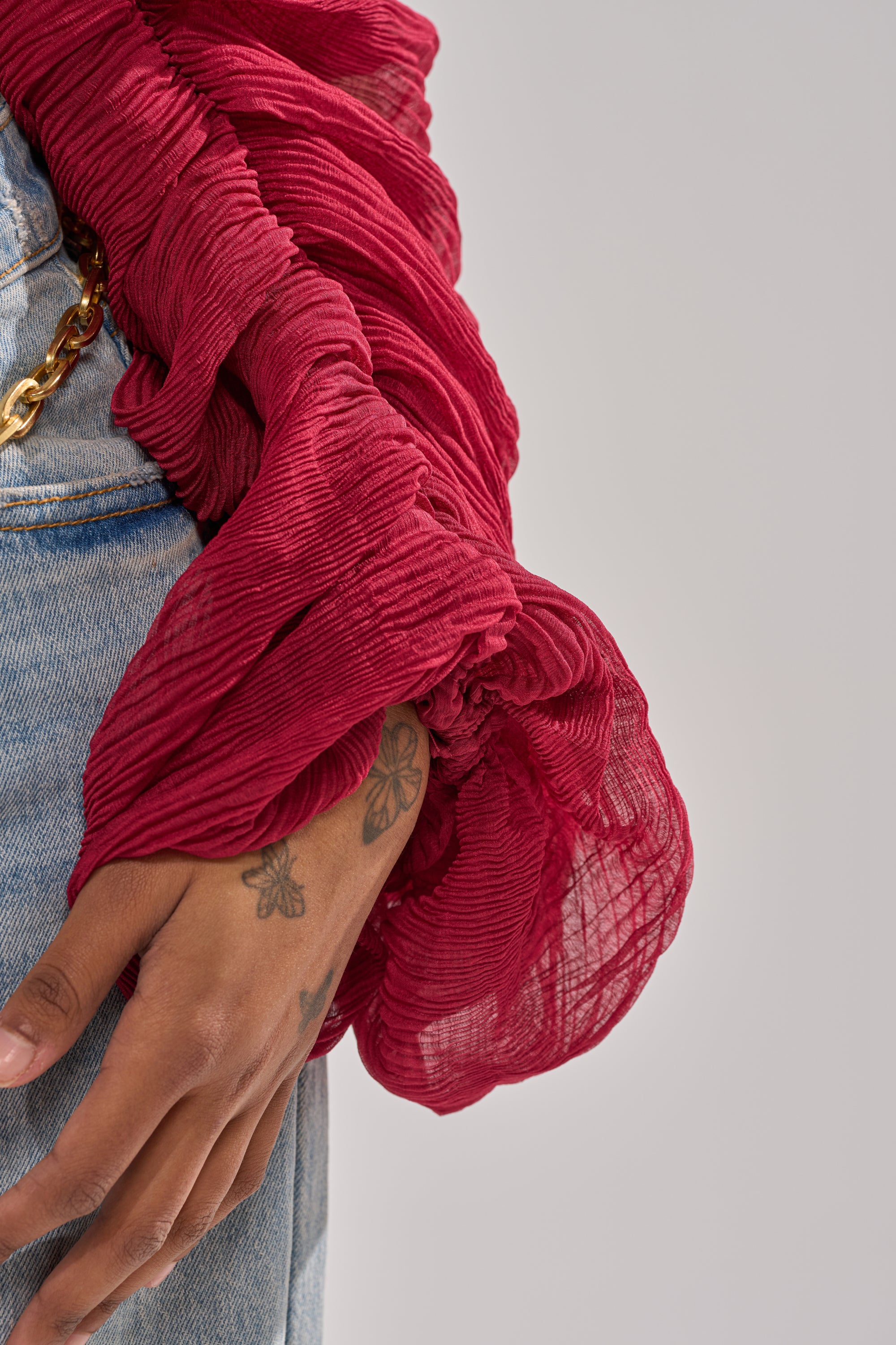 A close-up of a hand with butterfly tattoos wearing the GIRLY PLZ BLOUSE, a red sheer chiffon top with ruched balloon sleeves, paired with light blue jeans featuring a gold chain detail. The background is neutral gray. 7