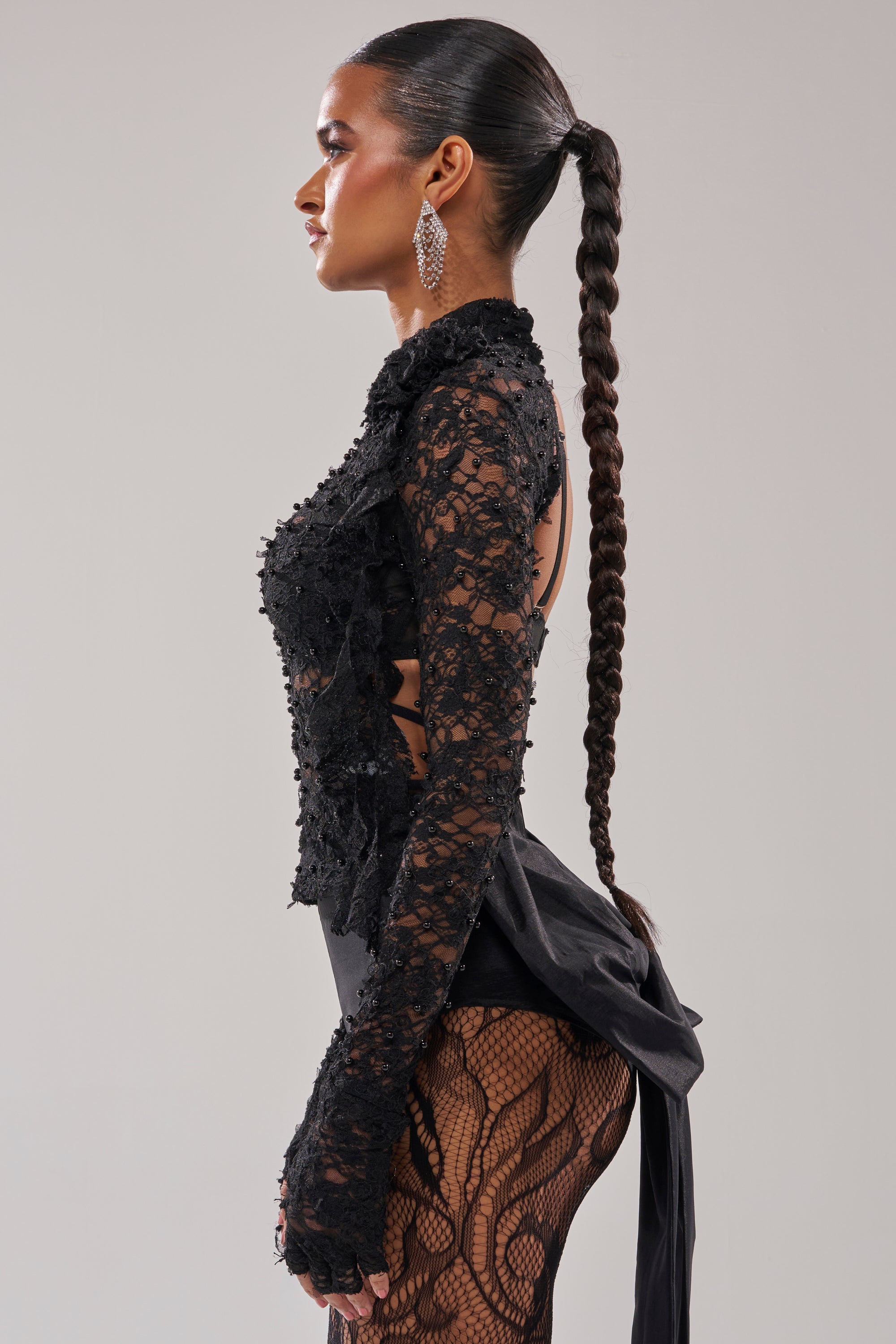 A woman with sleek, dark hair in a long braided ponytail models the LACED IN LOVE BLOUSE—a sheer floral lace top with cutout details—paired with black fishnet stockings and statement earrings, posing in profile against a light background.
