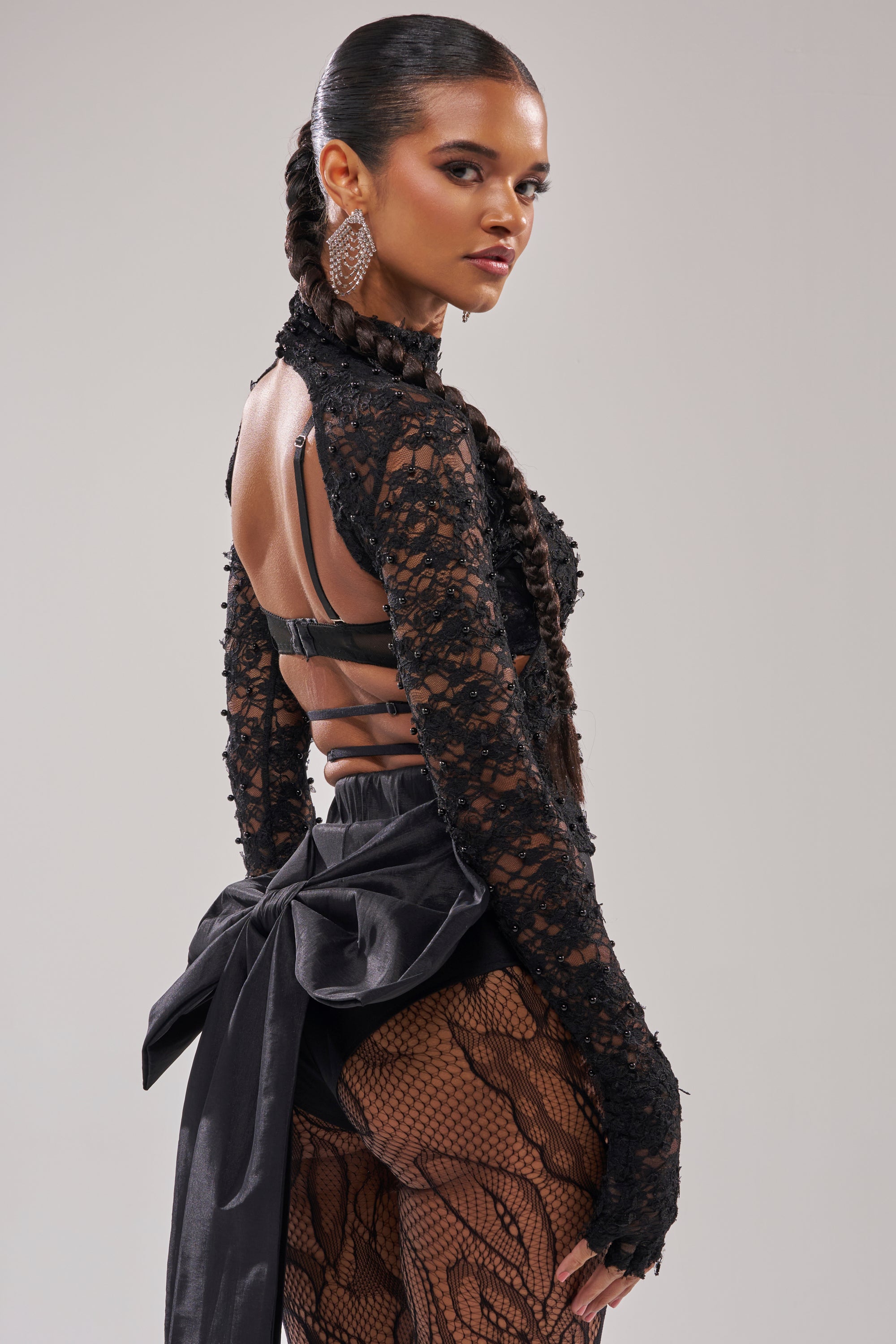 A woman with long, braided hair poses confidently in the LACED IN LOVE BLOUSE— a sheer floral lace top with cutouts—paired with dramatic earrings and fishnet stockings, standing against a plain light background and looking over her shoulder.
