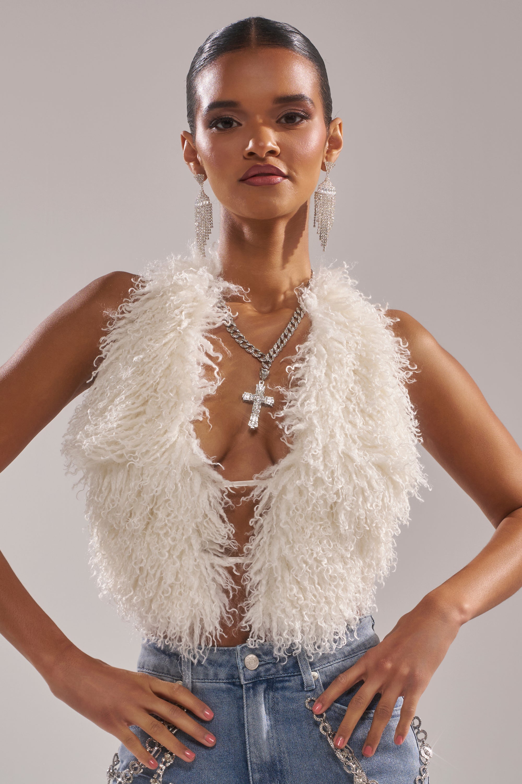 A woman stands confidently in the ACT UP FUR BLOUSE IN WHITE, styled with layered necklaces, large earrings, high-waisted blue jeans, slicked-back hair, and bold makeup.