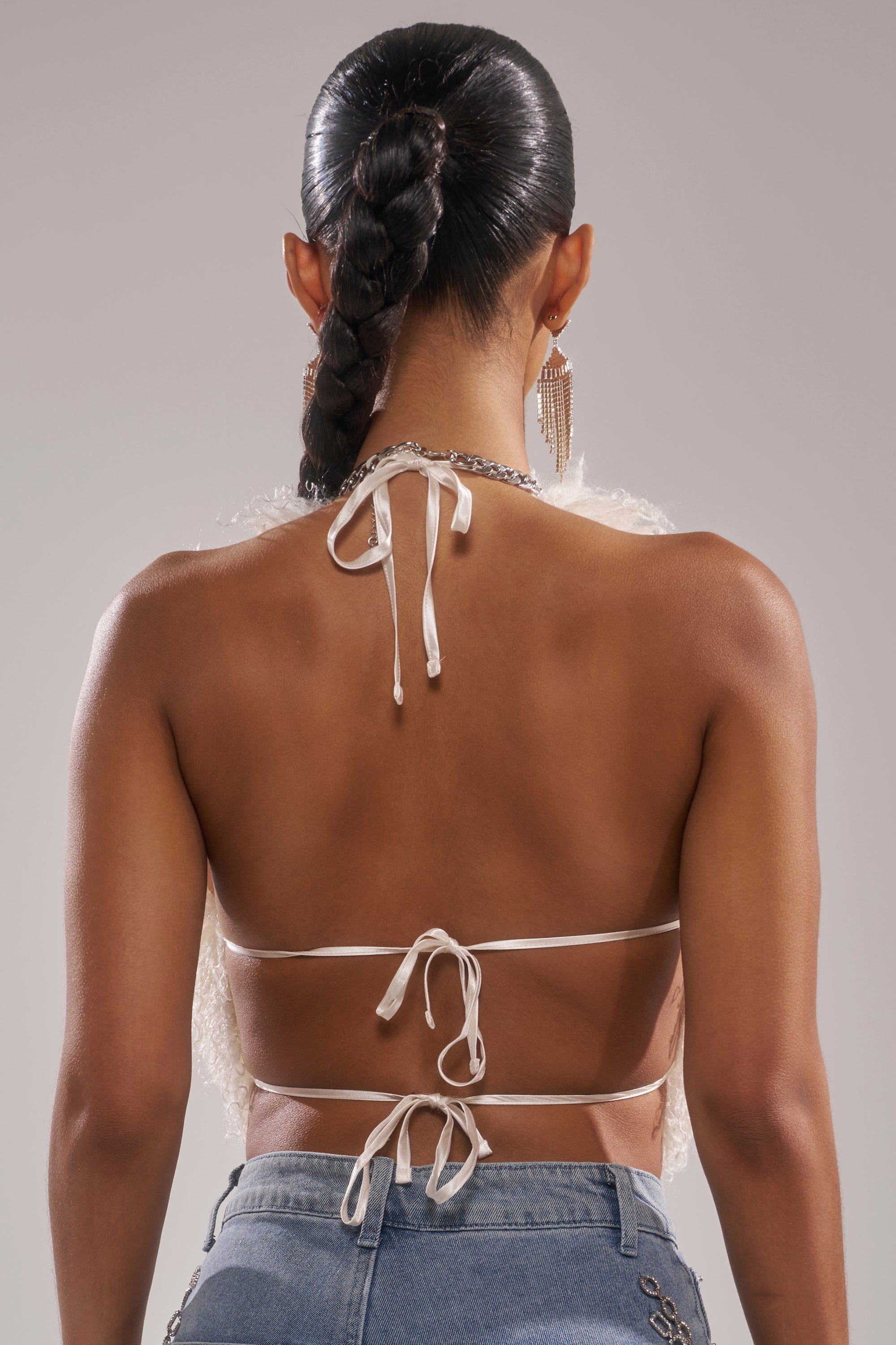 A person with braided hair is seen from behind in the ACT UP FUR BLOUSE IN WHITE, featuring an open-back tied with strings, paired with light blue jeans and large dangling earrings against a neutral background.