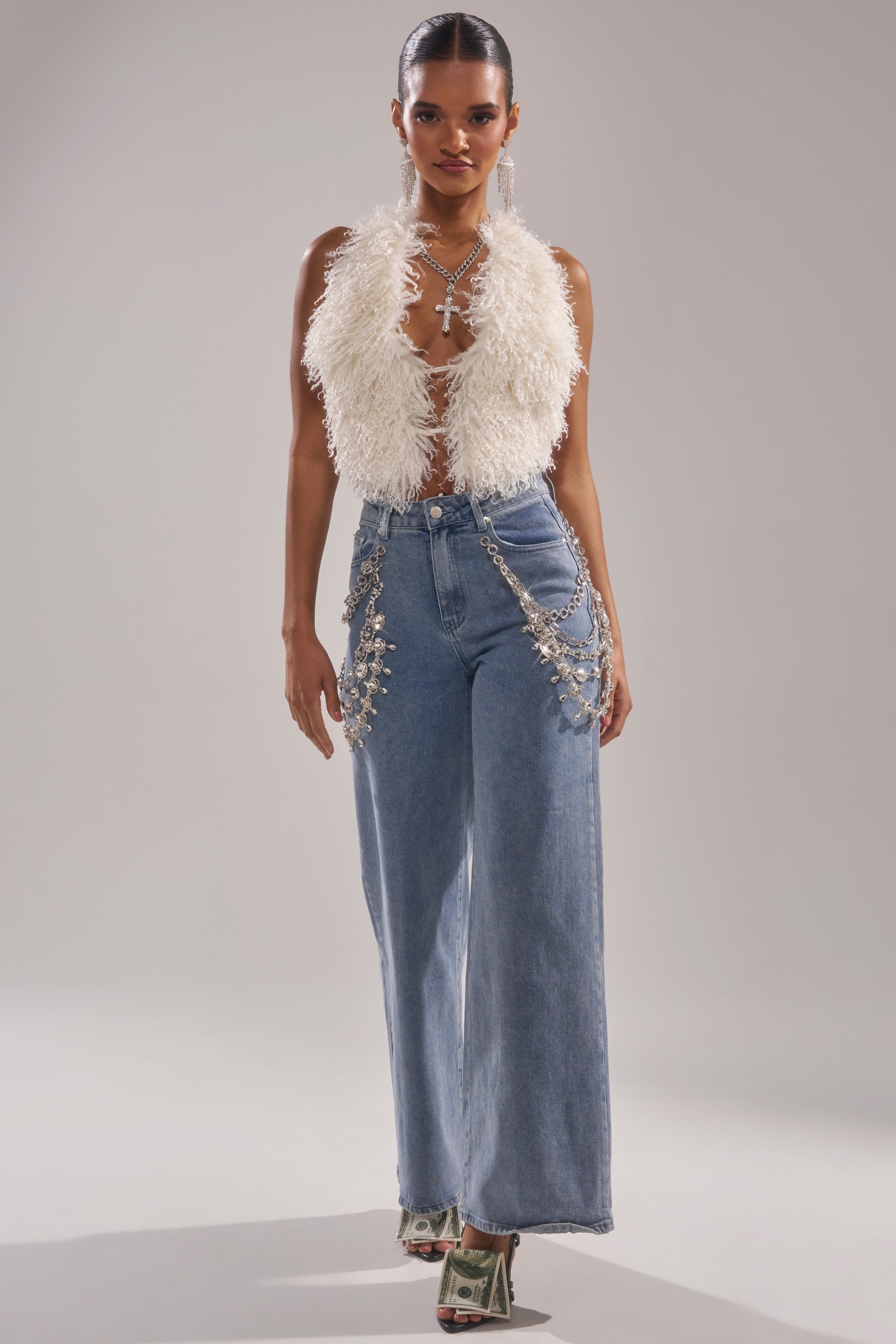 A woman poses confidently against a plain background in the ACT UP FUR BLOUSE IN WHITE, high-waisted wide-leg jeans with silver chain accents, and heels wrapped in dollar bills.