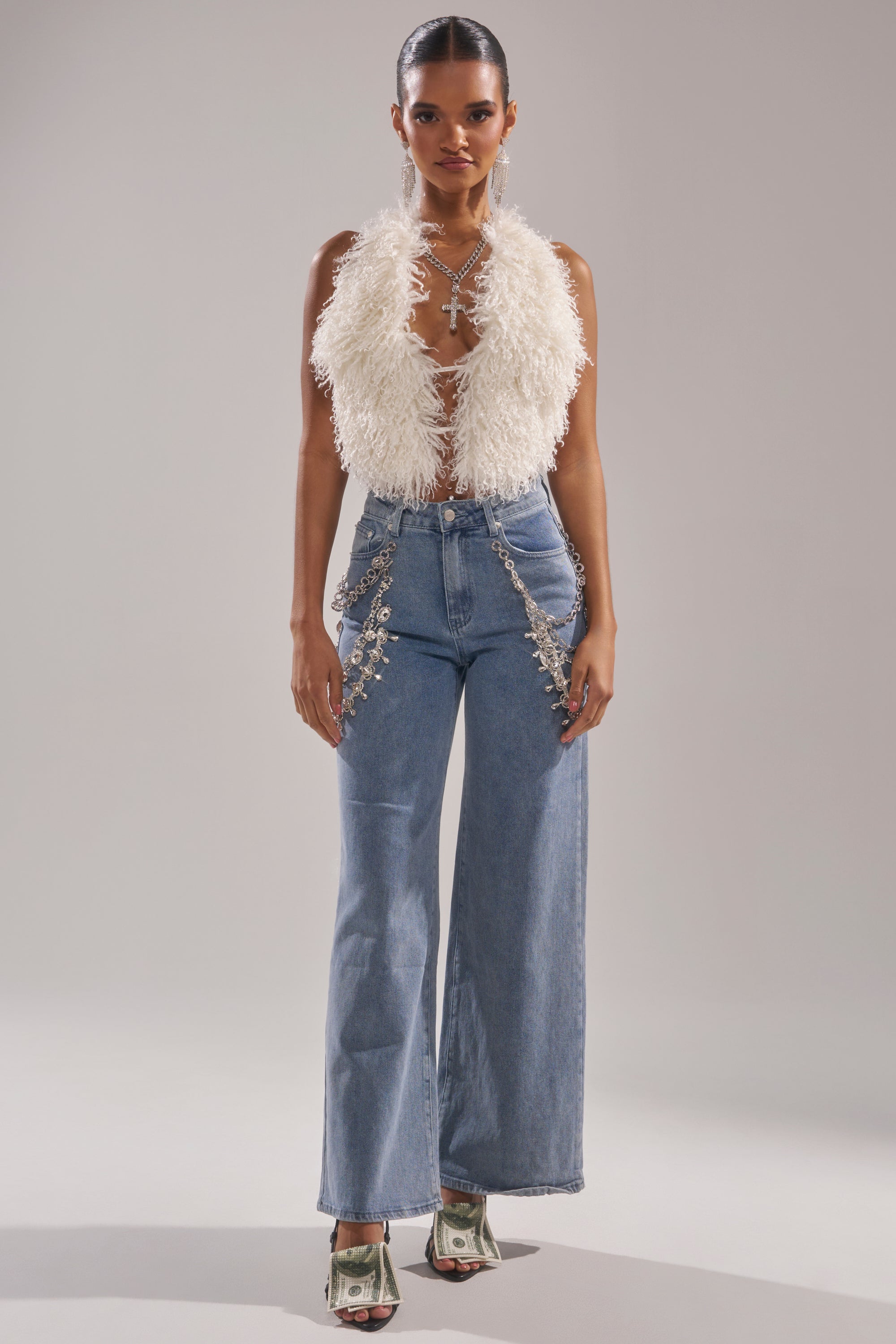 A woman stands facing forward wearing the ACT UP FUR BLOUSE IN WHITE, high-waisted blue jeans with rhinestone chains, and high-heeled sandals wrapped with dollar bills against a plain neutral background.