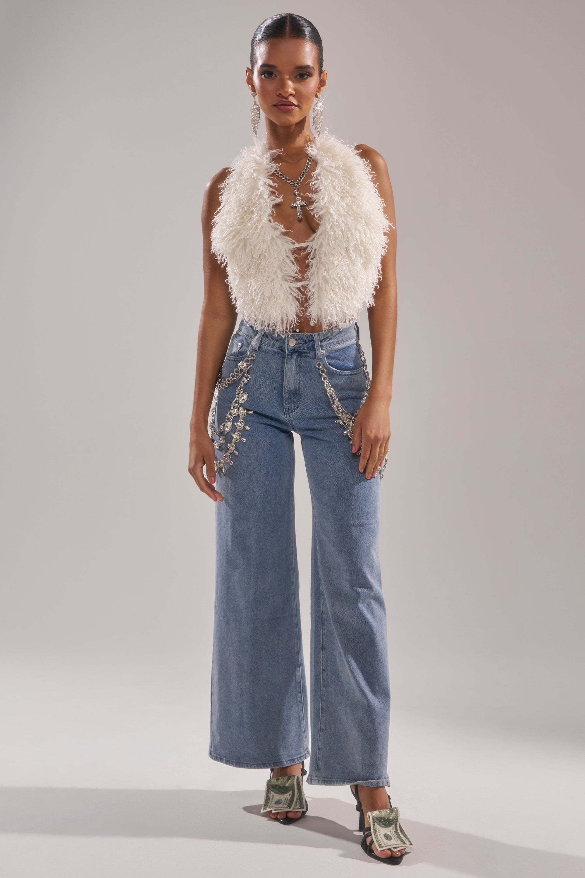 A woman wears the ACT UP FUR BLOUSE IN WHITE, a sleeveless cropped faux fur top, styled with light blue high-waisted wide-leg jeans, a jeweled chain belt, layered necklaces, and dollar bill heeled sandals.