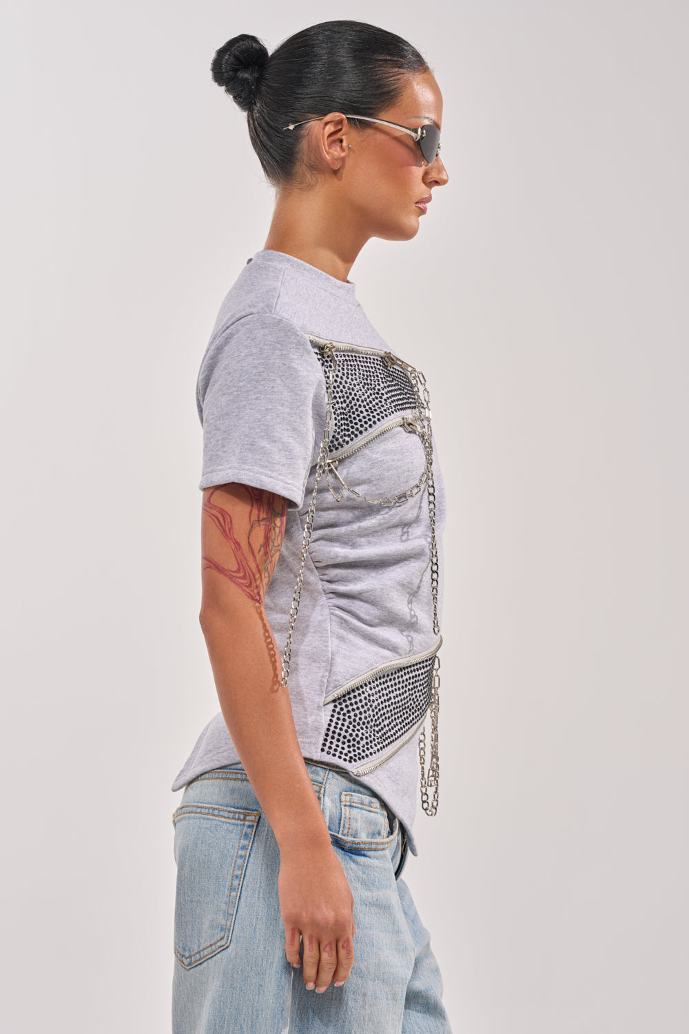 A person with dark hair in a bun, sunglasses, light blue jeans, and a visible arm tattoo stands in profile against a plain background, wearing the DONT MESS OVERSIZED SWEATSHIRT. 3