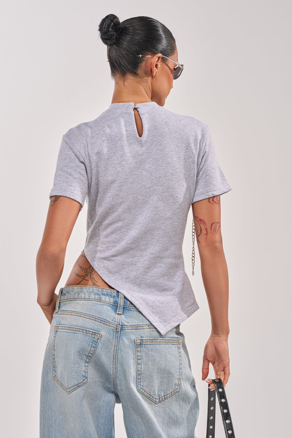 A person with dark hair in a bun, wearing sunglasses and the DONT MESS OVERSIZED SWEATSHIRT in light gray streetwear style with short sleeves, plus light blue jeans, stands facing away, tattoos visible on their lower back and hip. 4