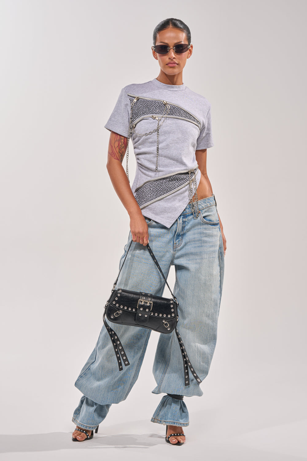 Wearing the DONT MESS OVERSIZED SWEATSHIRT with chain details, a person in sunglasses pairs it with baggy light blue jeans, heeled sandals, and a black studded handbag, standing confidently against a plain background. 5
