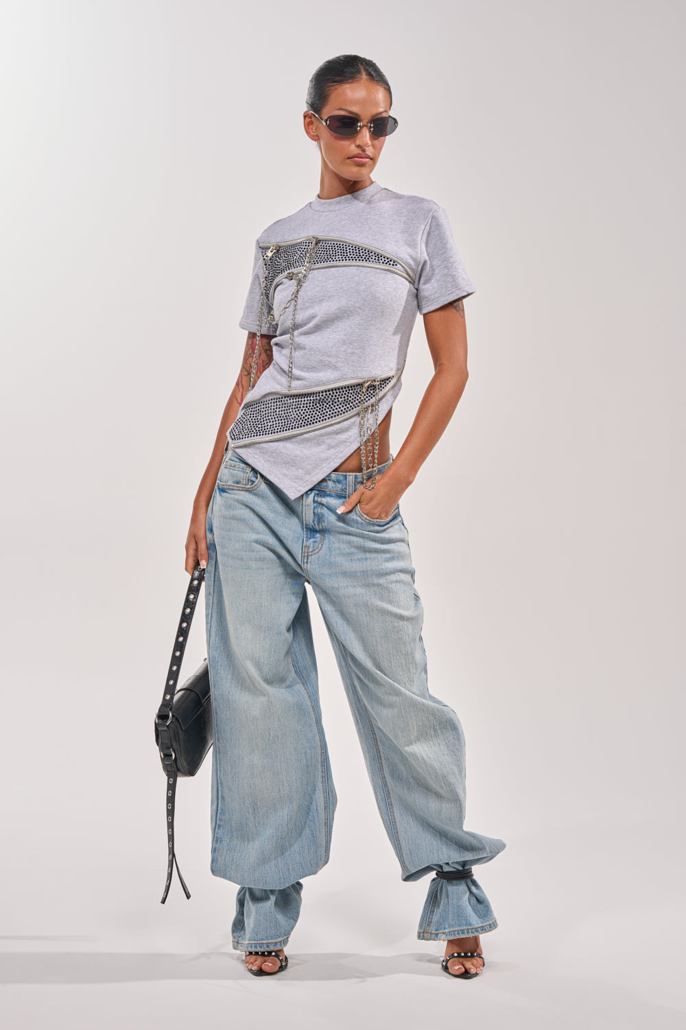 A woman poses in sunglasses, the DONT MESS OVERSIZED SWEATSHIRT with short sleeves and rhinestone chains, loose light blue baggy jeans, and heels. She holds a black handbag and has her hair pulled back. 6