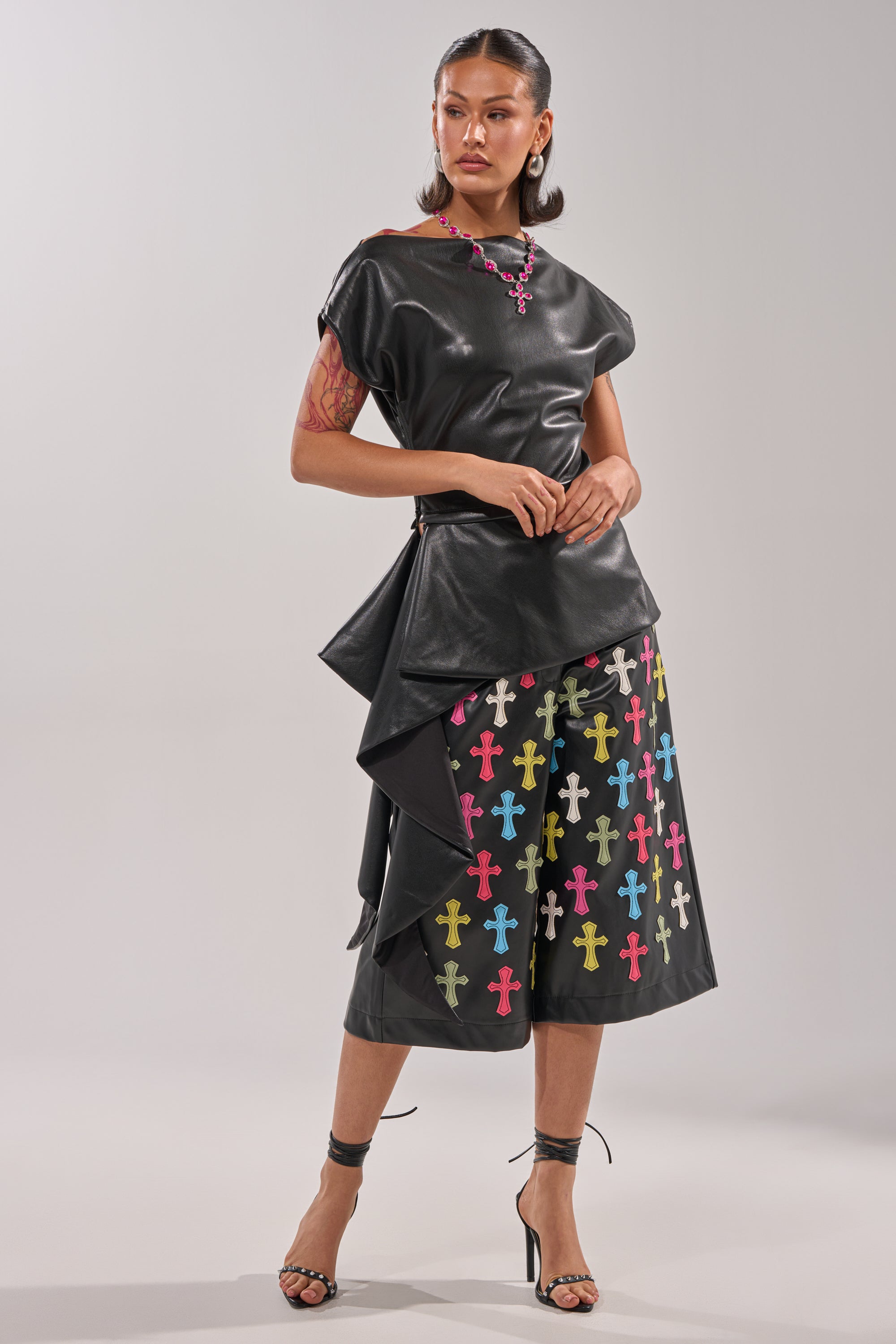 A woman wears the ALEXA FAUX LEATHER BLOUSE with an asymmetrical hem, paired with black culottes featuring colorful cross patterns. She accessorizes with hoop earrings, strappy heels, and has her hair slicked back. 8