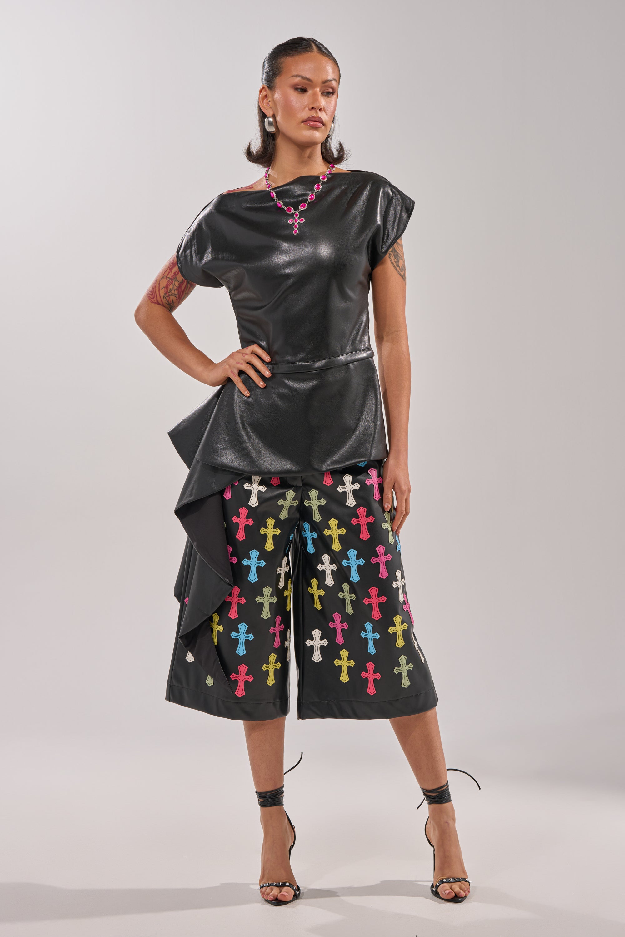 A woman stands confidently in the ALEXA FAUX LEATHER BLOUSE—a black peplum top with side detail—paired with black culottes featuring colorful cross patterns. She accessorizes with strappy heels, red lipstick, jewelry, and shows off her arm tattoos. 9