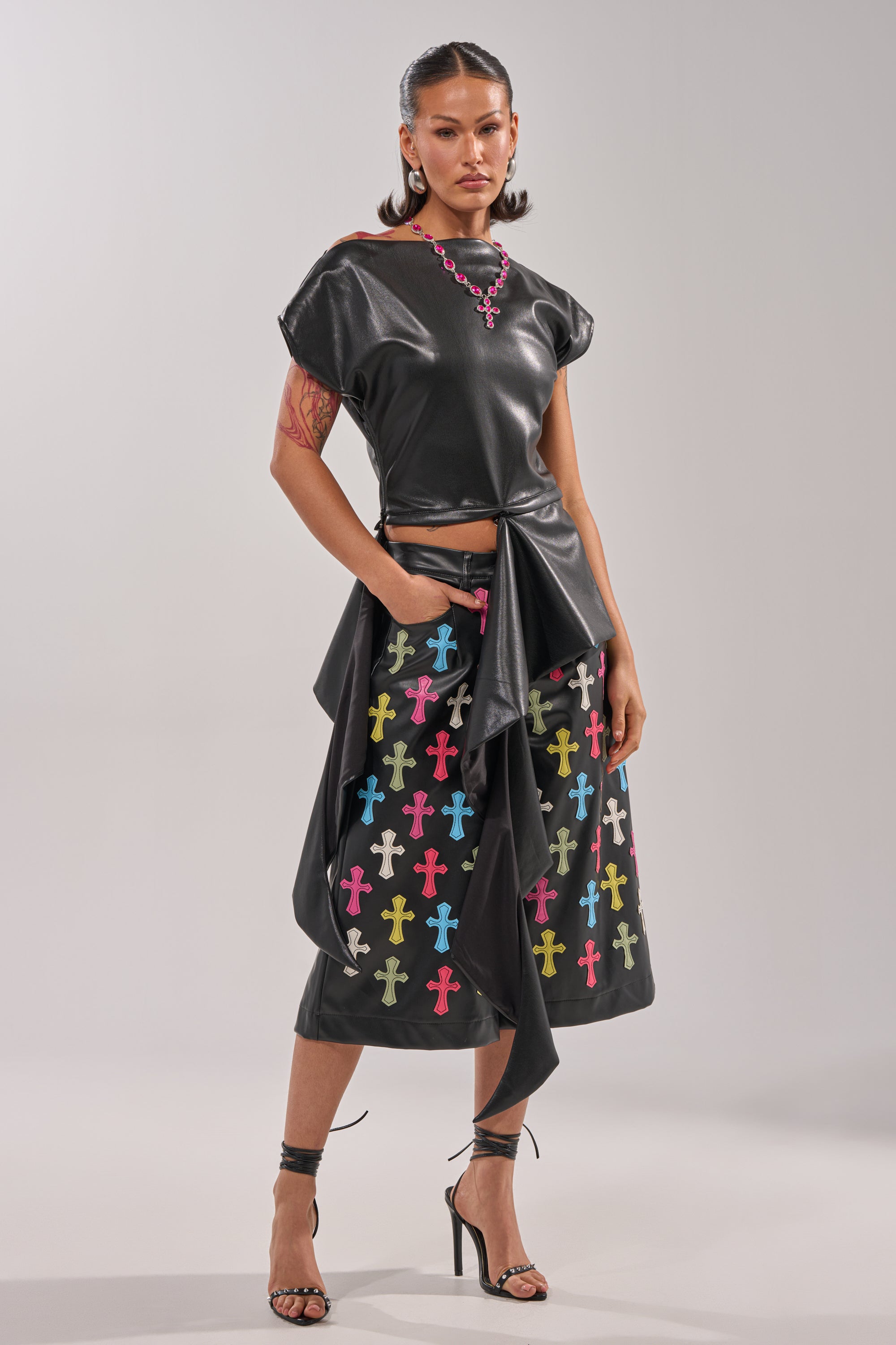 Wearing the ALEXA FAUX LEATHER BLOUSE and a matching skirt with colorful cross patterns, a woman in high heels, hoop earrings, and long pigtails strikes a confident pose against a neutral background. 10
