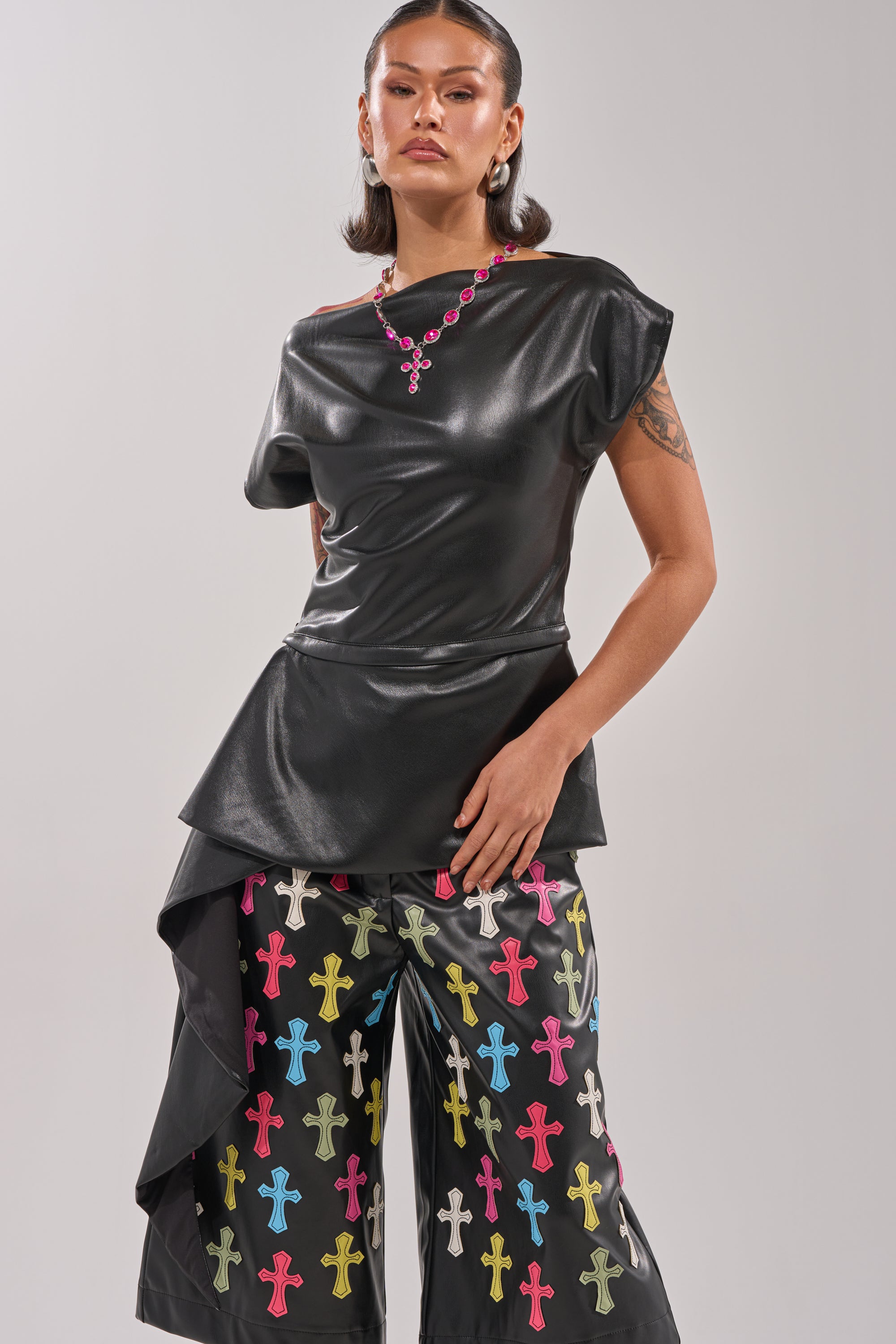Someone stands confidently in the ALEXA FAUX LEATHER BLOUSE, a black peplum top with an asymmetrical accent, paired with black pants featuring colorful cross patterns. They wear a pink necklace and show tattooed arms against a light gray background. 1