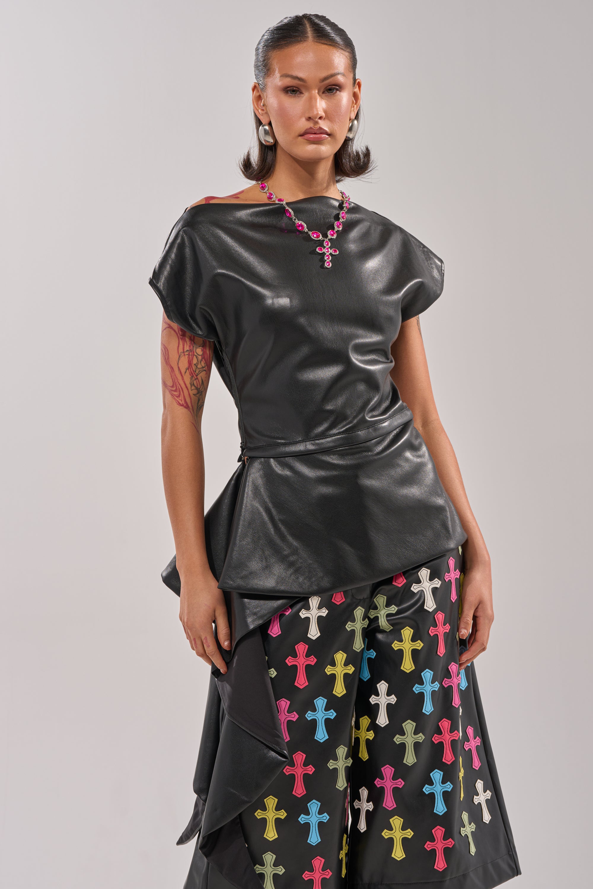A woman stands against a plain background in the ALEXA FAUX LEATHER BLOUSE, styled off-the-shoulder with wide-leg black pants featuring colorful cross patterns. Her sleek, shoulder-length hair is complemented by a pink cross necklace. 2