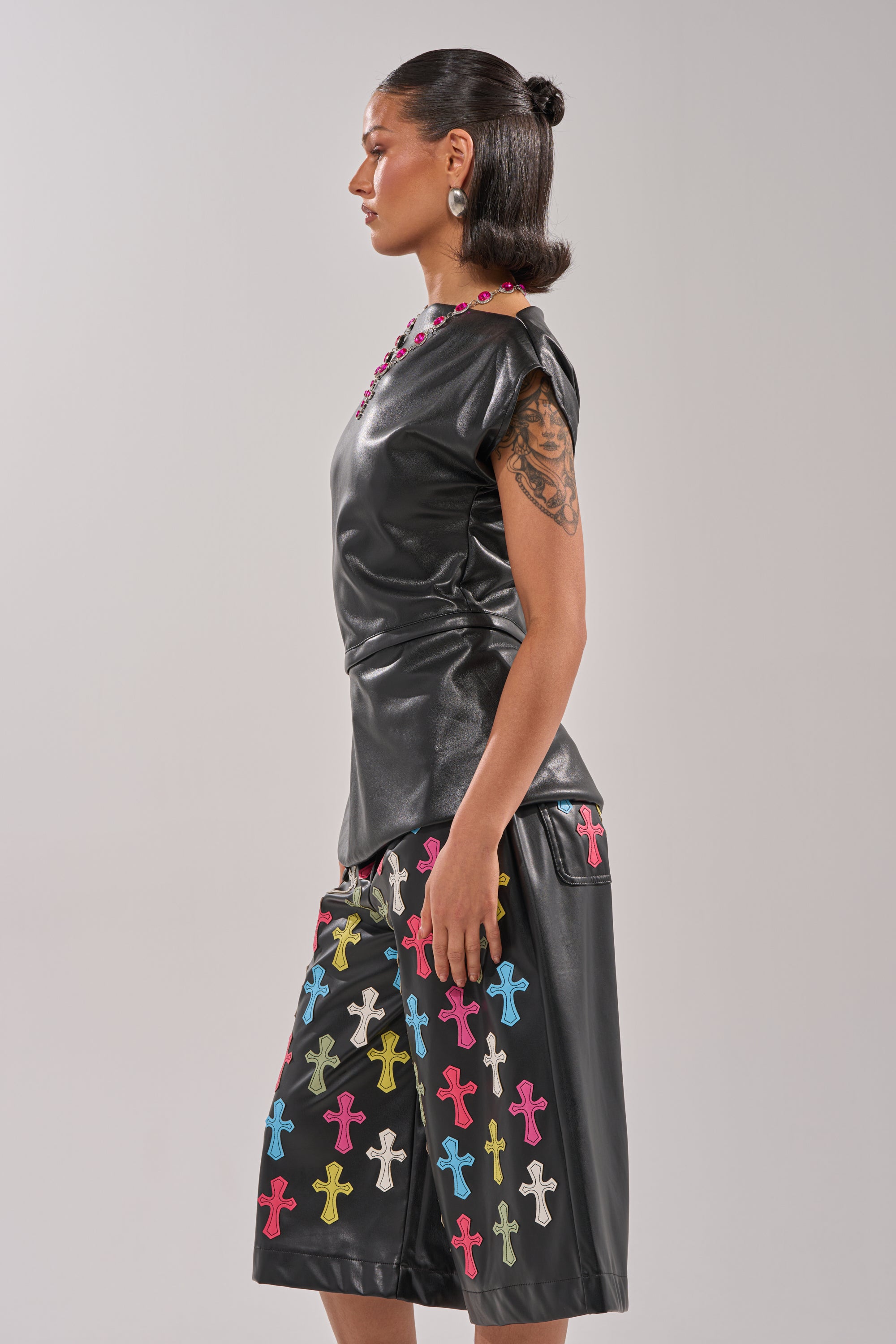 A woman in profile wears the ALEXA FAUX LEATHER BLOUSE and matching culottes with colorful cross patterns. She sports slicked-back hair, statement earrings, a visible arm tattoo, and poses against a plain light background. 4