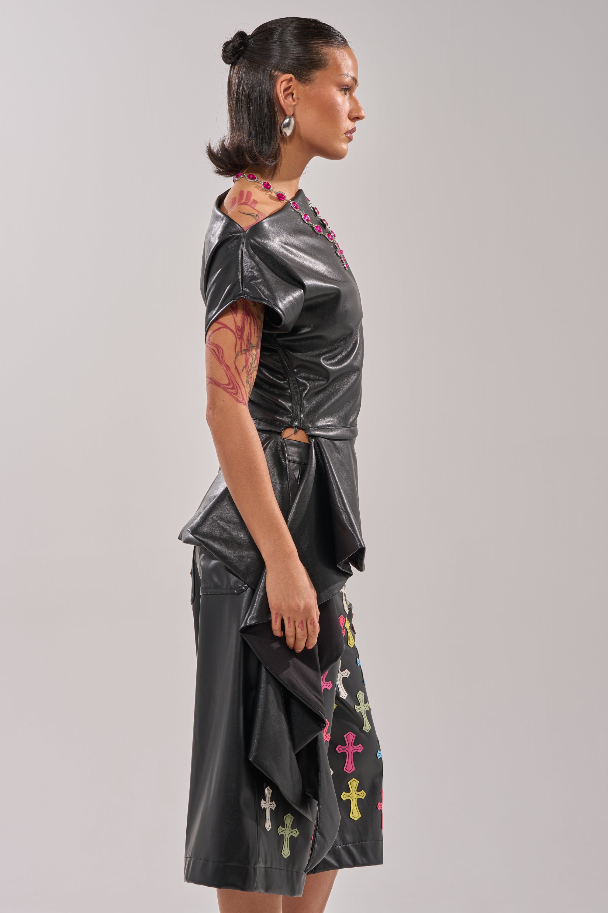 A woman stands in profile wearing the ALEXA FAUX LEATHER BLOUSE and a skirt with colorful crosses. She has slicked-back hair, large earrings, and visible arm tattoos. 5
