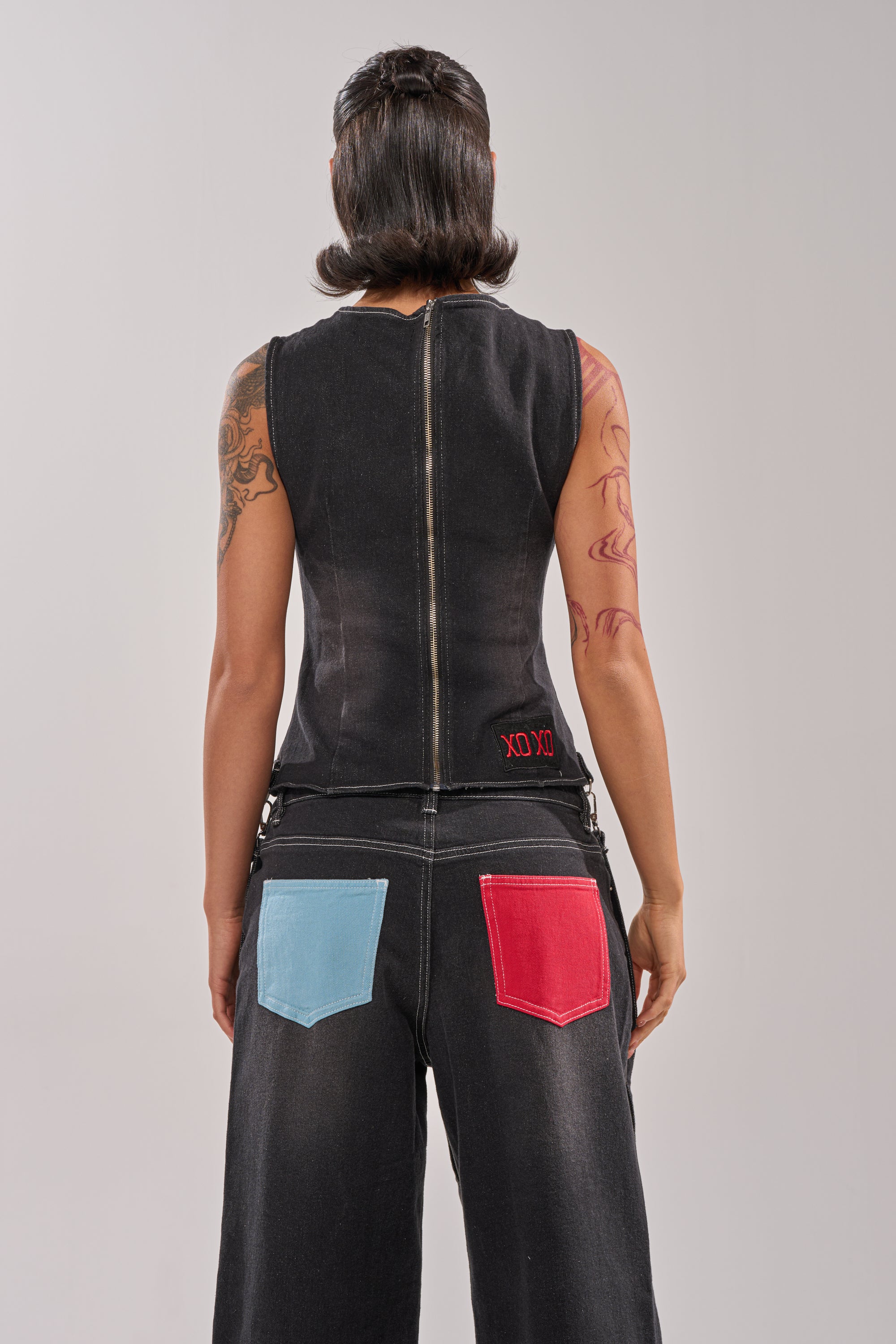 Wearing the ALEXA FAUX LEATHER BLOUSE, a person with short dark hair and arm tattoos stands facing away, showing off an off-the-shoulder style with a back zipper, paired with black jeans with blue and red back pockets. 6