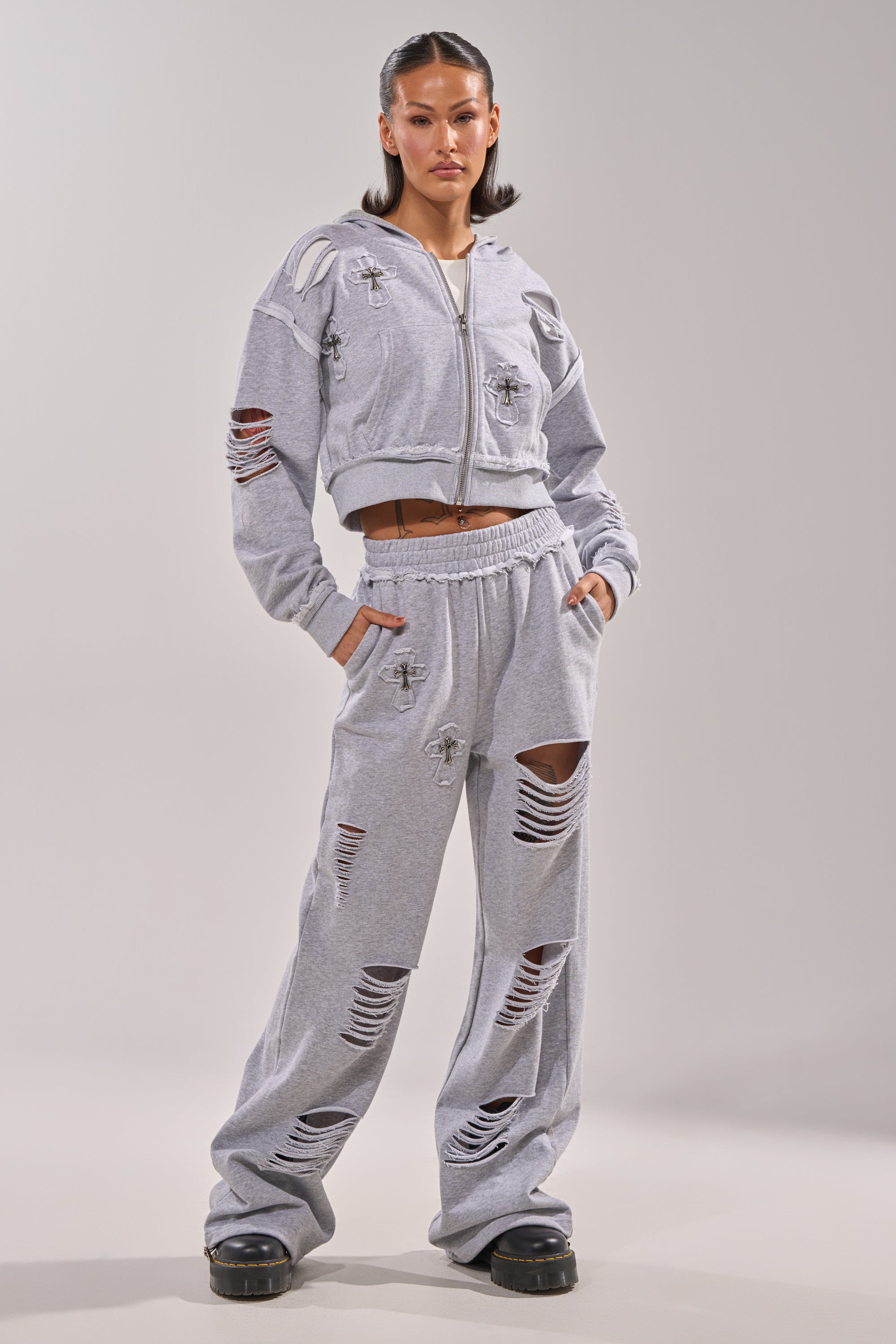 A woman stands confidently in the CROSSED OUT DISTRESSED WIDE LEG JOGGER and a matching ripped jacket, paired with black chunky shoes, hands in her pockets against a plain background. 0