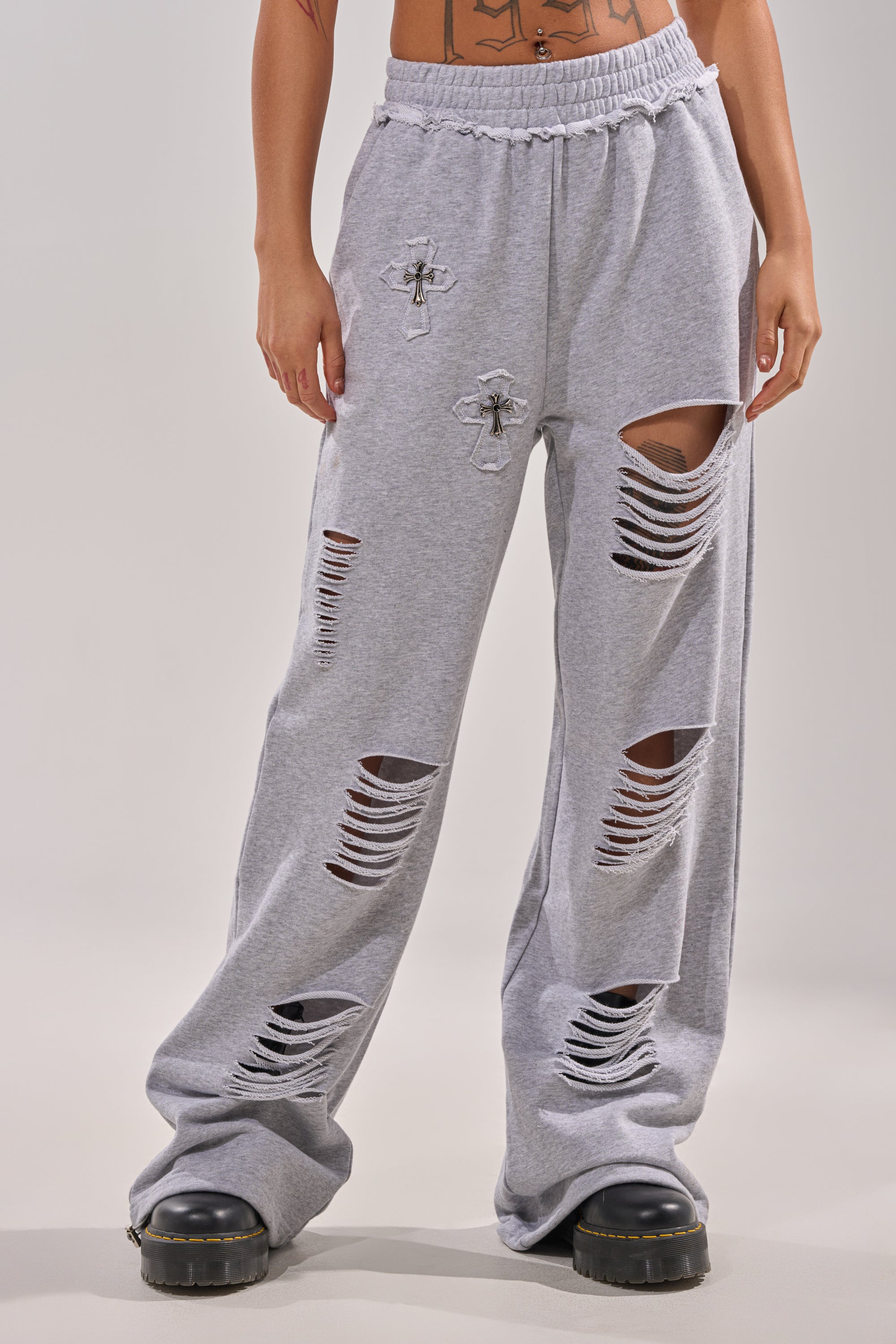 A person stands against a plain background wearing the CROSSED OUT DISTRESSED WIDE LEG JOGGER, featuring light gray fabric, large ripped sections, and cross patches, paired with black chunky shoes. 1