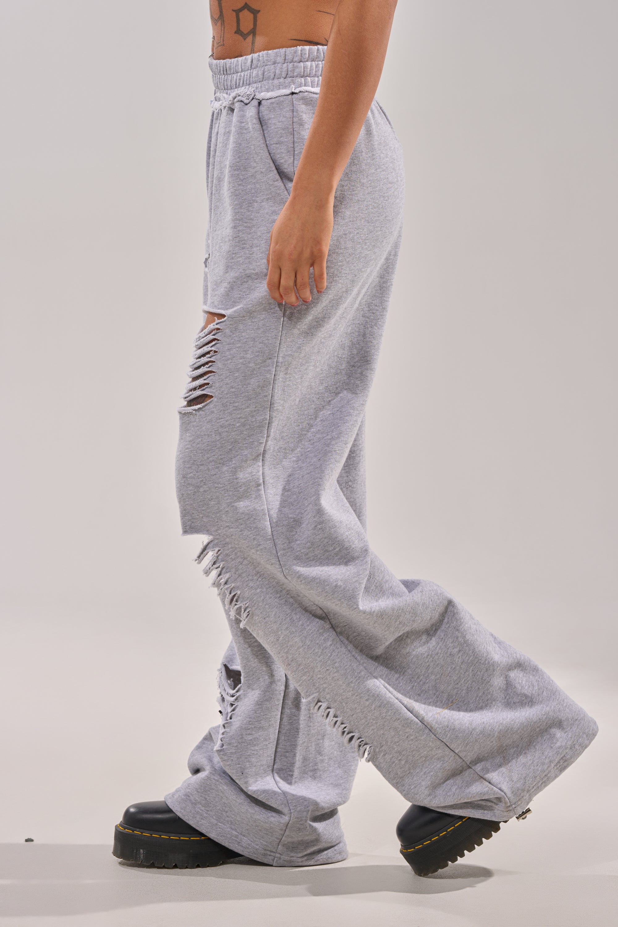 Someone stands against a plain light background wearing the CROSSED OUT DISTRESSED WIDE LEG JOGGER in light gray with ripped thigh and knee details, paired with black chunky platform shoes. 2