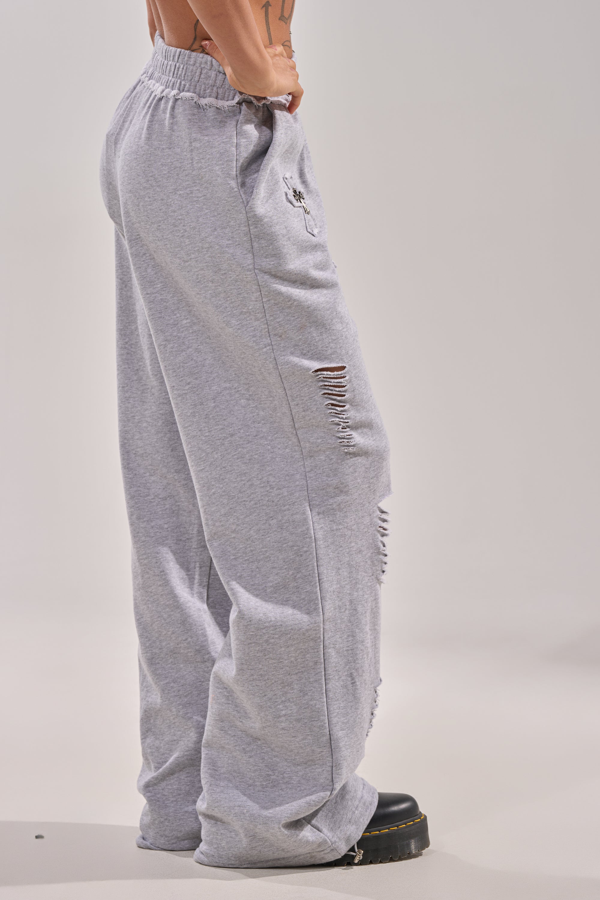Model wears the CROSSED OUT DISTRESSED WIDE LEG JOGGER—loose-fit, light gray joggers featuring horizontal thigh and knee rips, elastic waistband, and paired with black chunky shoes. Plain light gray background. 3