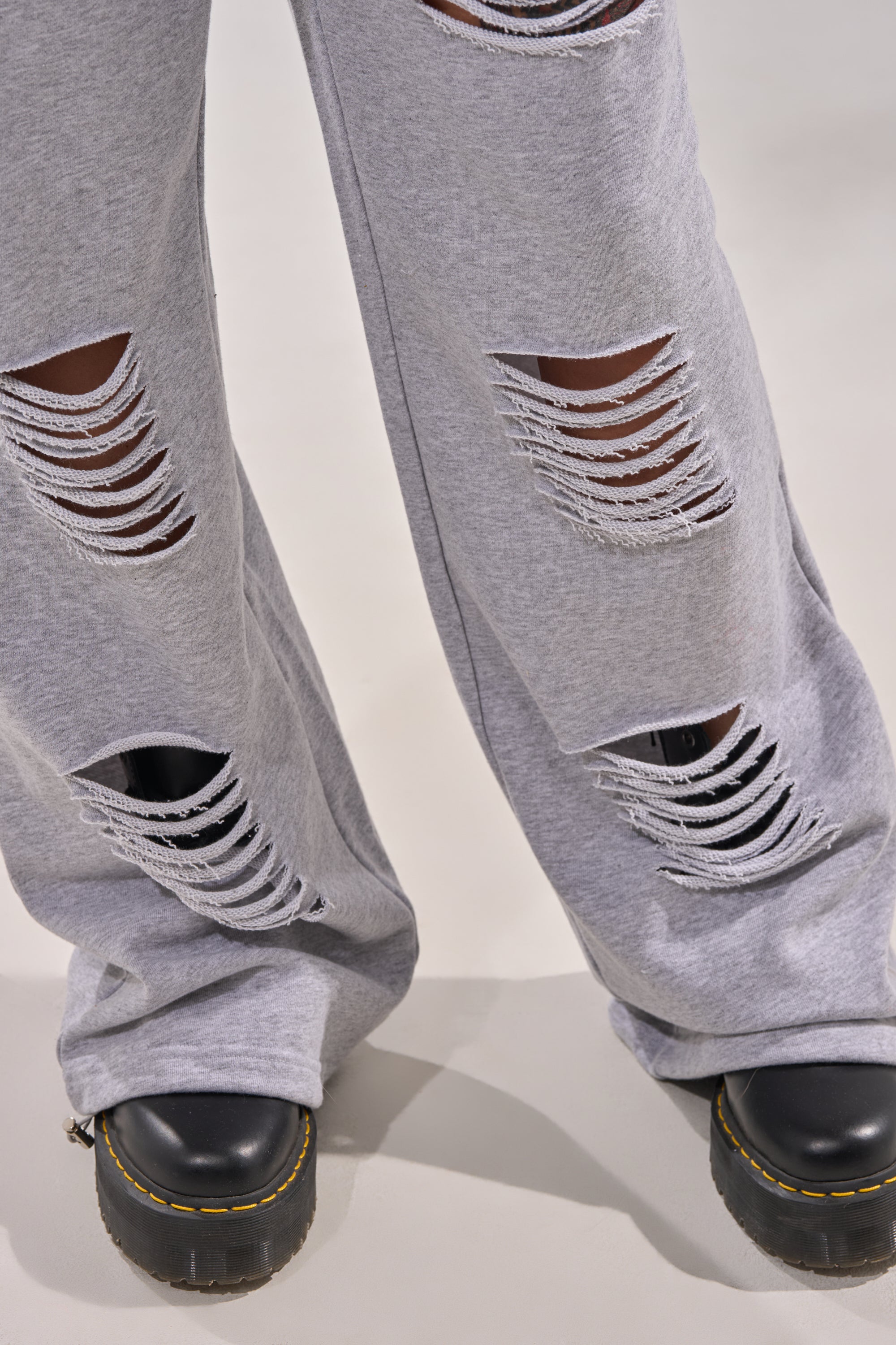 Close-up of a person wearing the CROSSED OUT DISTRESSED WIDE LEG JOGGER in light gray with large horizontal rips, paired with black platform shoes featuring yellow stitching, set against a light background. 6