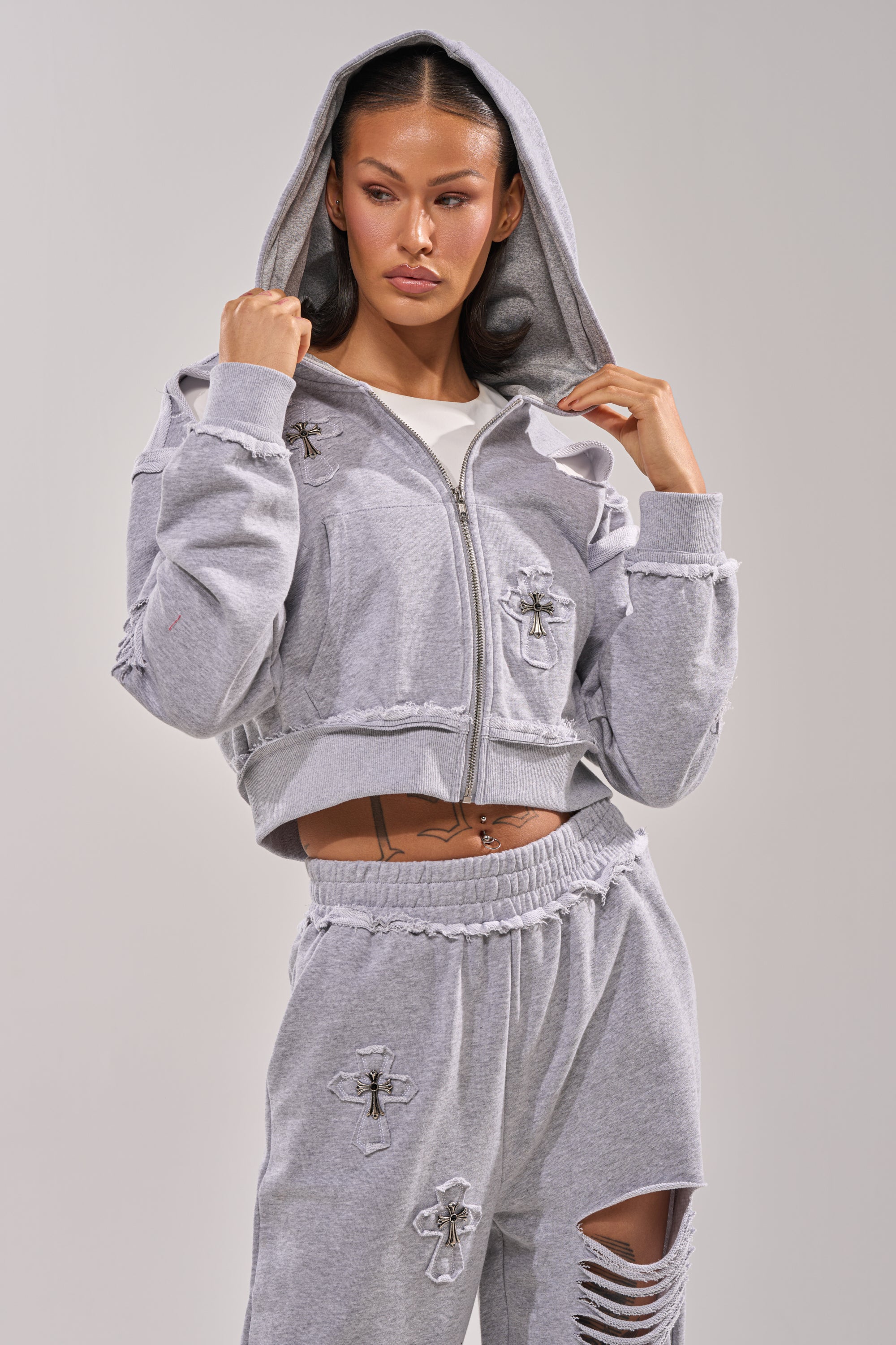 A woman poses confidently in the CROSSED OUT DISTRESSED ZIP UP SWEATSHIRT, a light gray hoodie with cross patches, distressed details, and silver cross pendants. She pairs it with matching elastic-waist pants and holds the collar with the hood up. 0