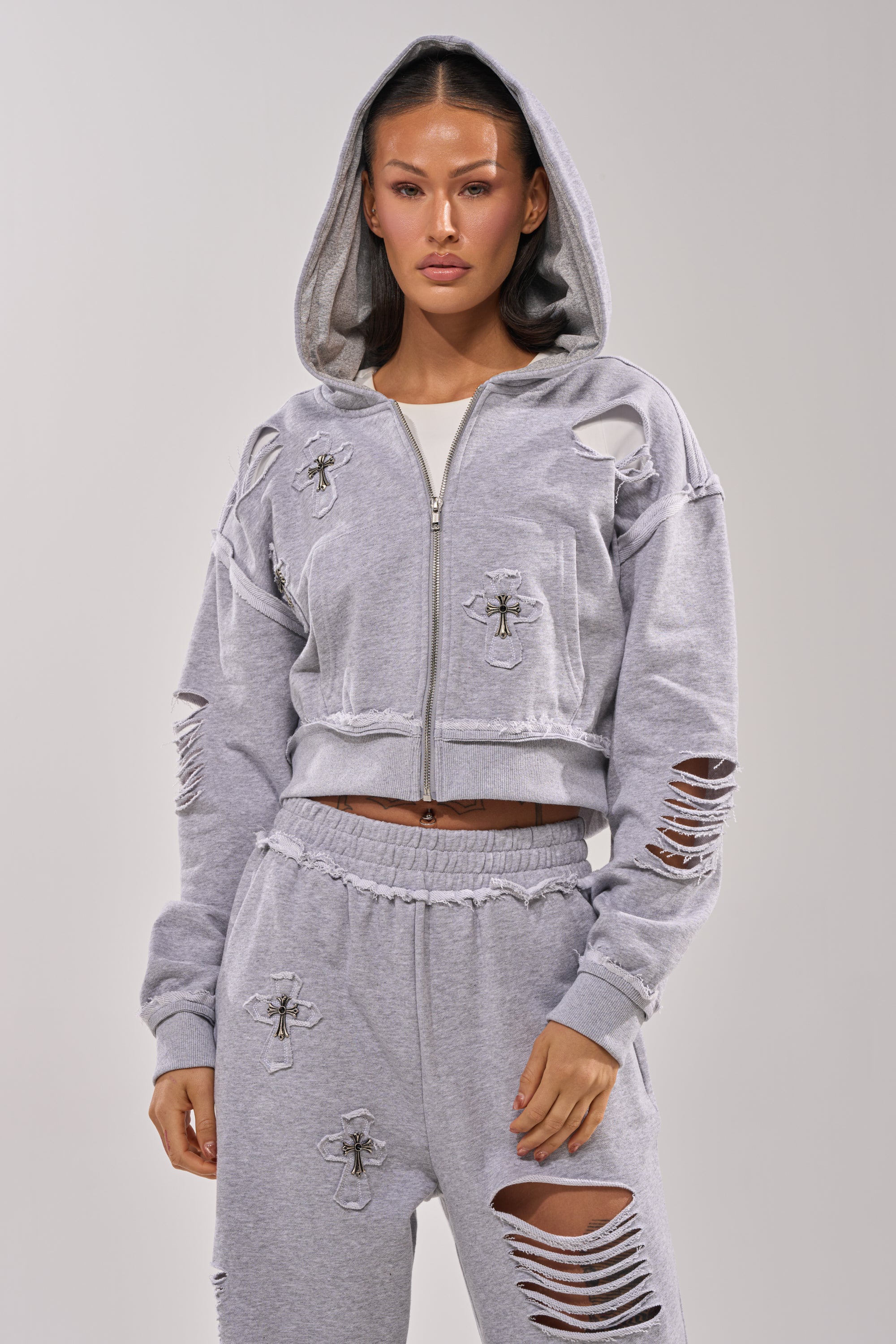 A woman wears the CROSSED OUT DISTRESSED ZIP UP SWEATSHIRT, a light gray hoodie with ripped details and cross patches, unzipped and hood up, styled with silver cross pendants against a plain light background. 1
