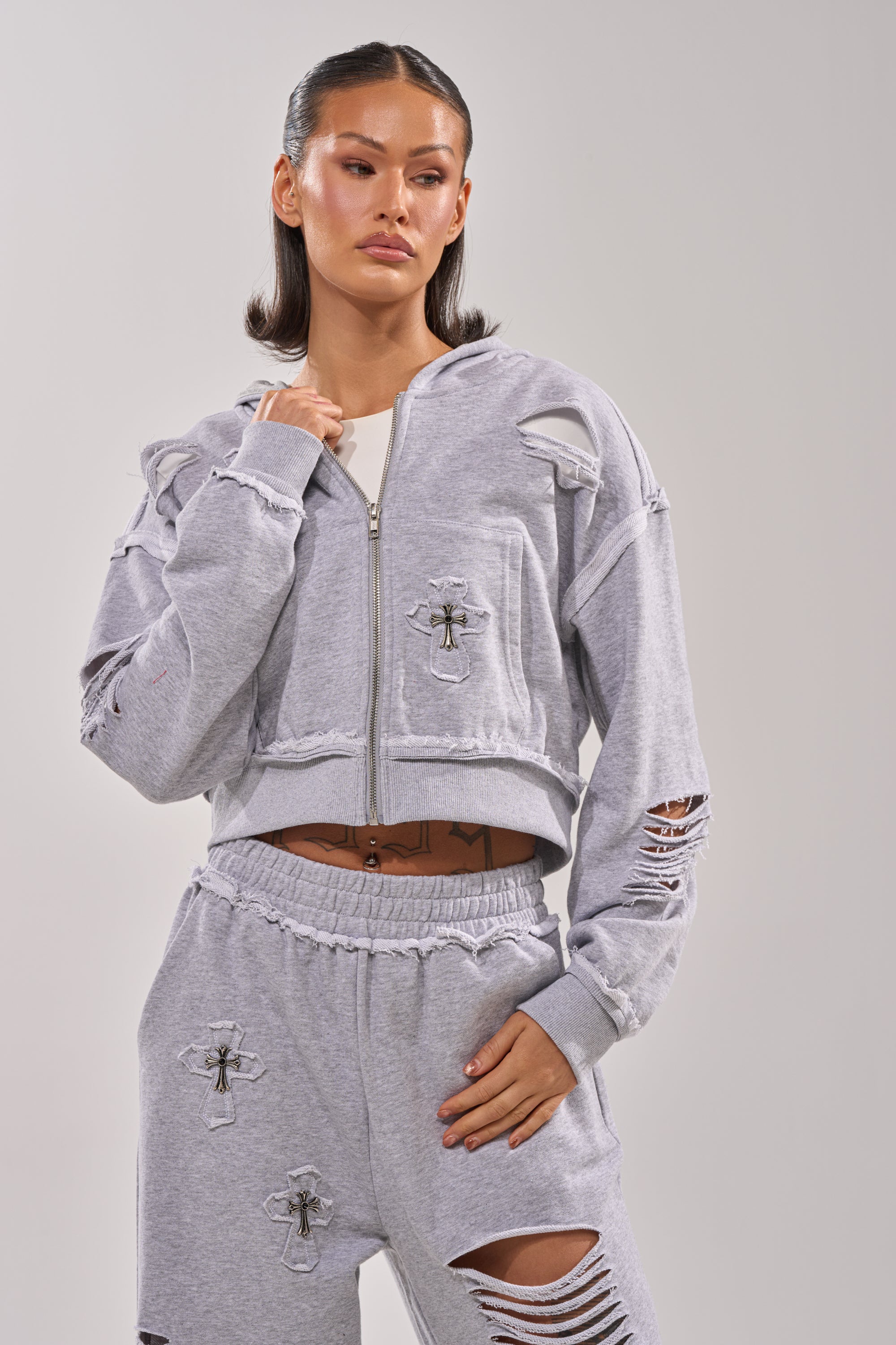 A woman with straight dark hair and a neutral expression wears the CROSSED OUT DISTRESSED ZIP UP SWEATSHIRT in light gray, featuring cross patches, paired with matching sweatpants. She stands against a plain background, holding the collar. 2