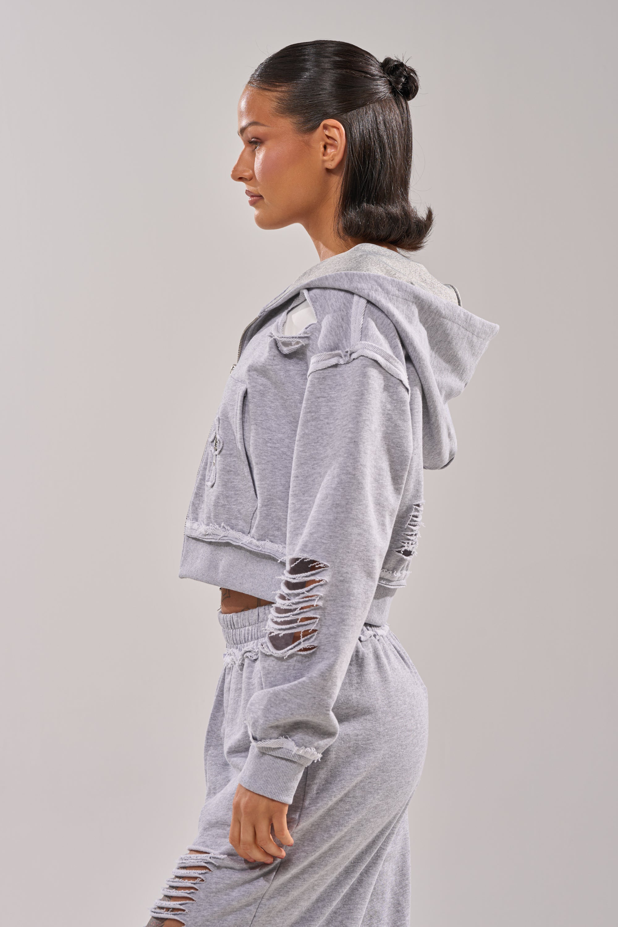 A woman with slicked-back hair models the CROSSED OUT DISTRESSED ZIP UP SWEATSHIRT in light gray, featuring a cropped fit, rips, a hood, and matching sweatpants, standing in profile against a plain background. 3
