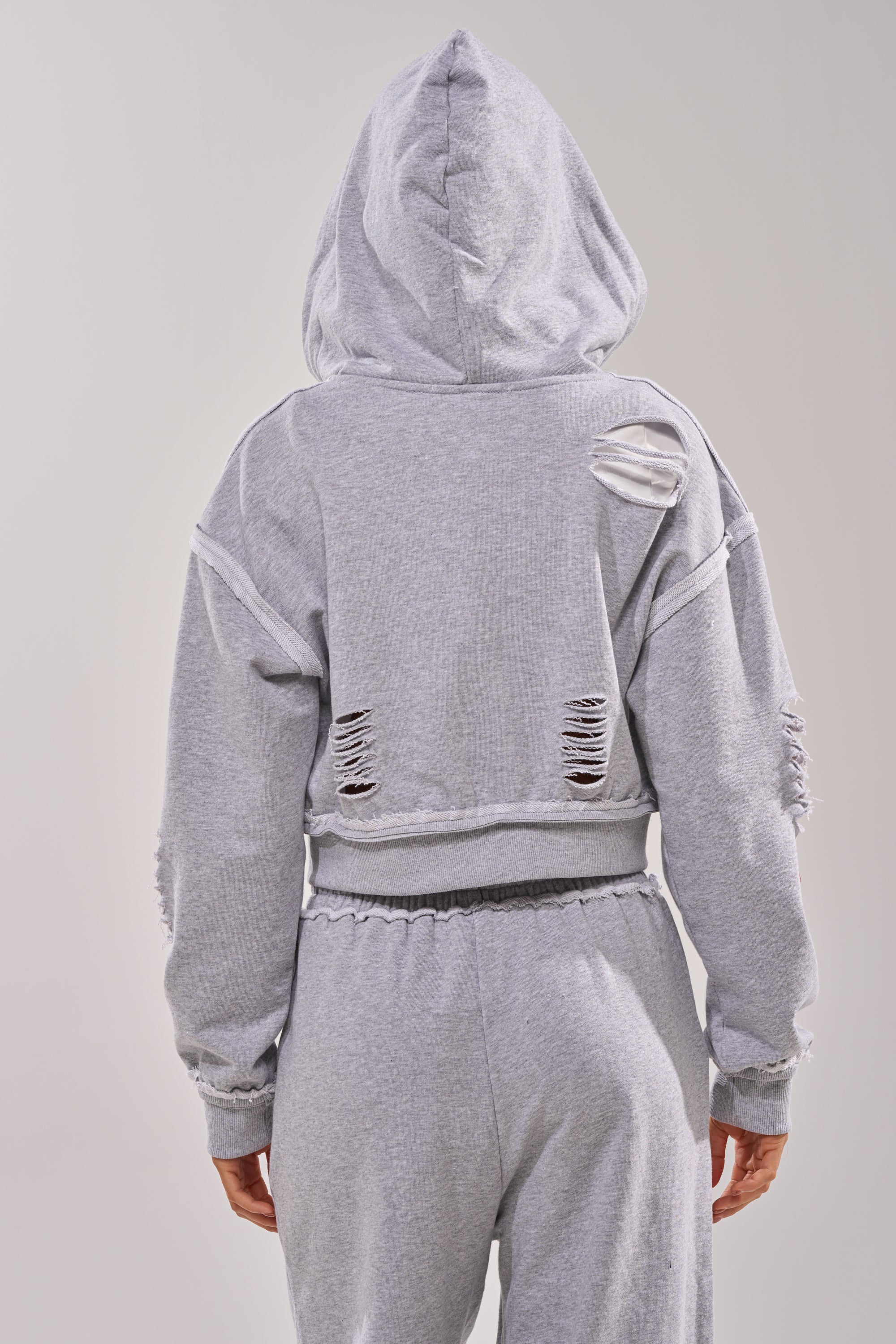 A person is seen from the back wearing the CROSSED OUT DISTRESSED ZIP UP SWEATSHIRT in light gray with matching sweatpants, featuring intentional rips and frayed details on the back and sleeves for a trendy, relaxed look. 4