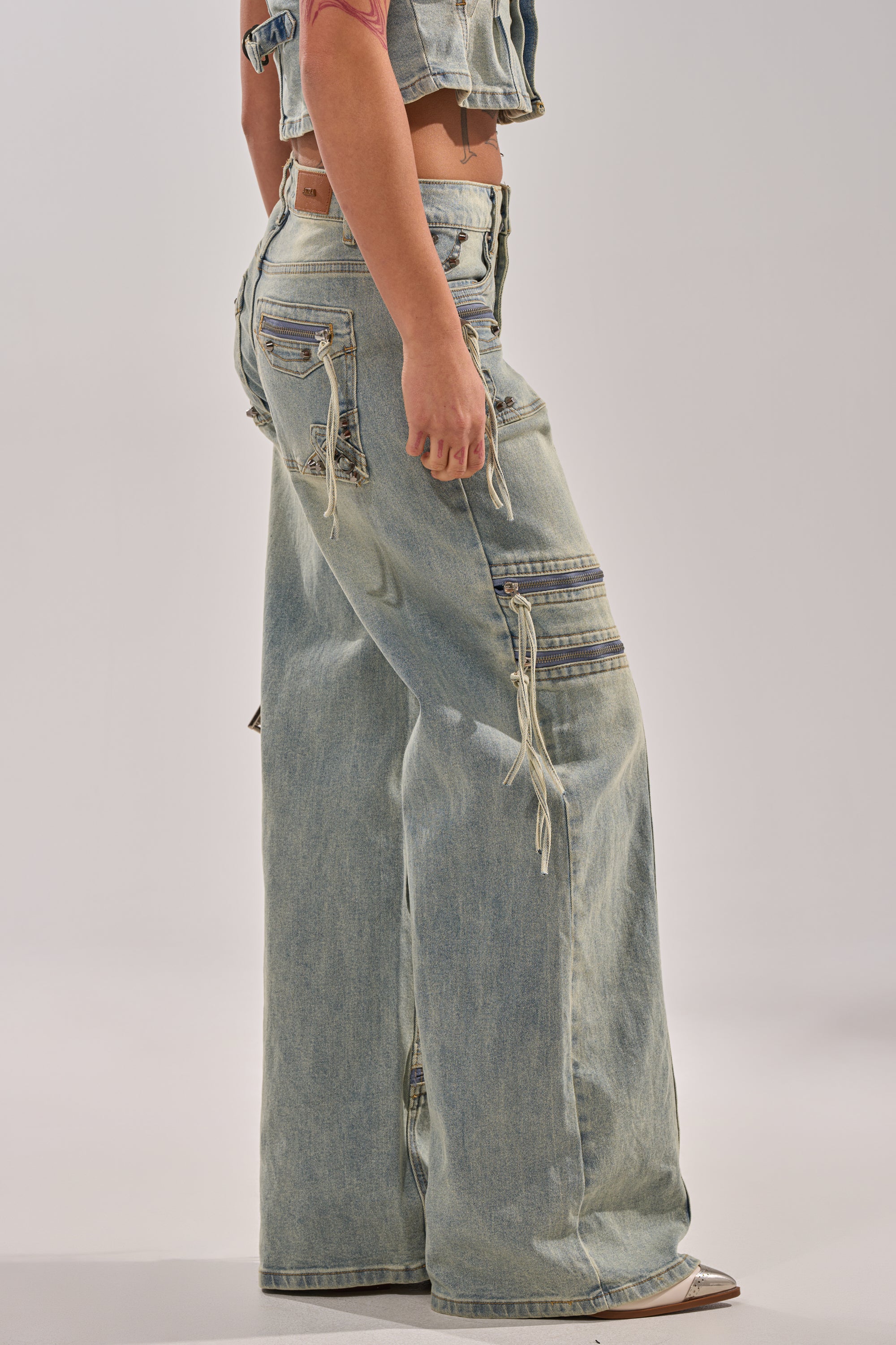 A person stands sideways against a plain background, showing only their lower body in the COMEBACK ERA WIDE LEG DENIM JEANS—light blue, wide-leg denim with utility pockets and zippers that capture the essence of Y2K streetwear. 2