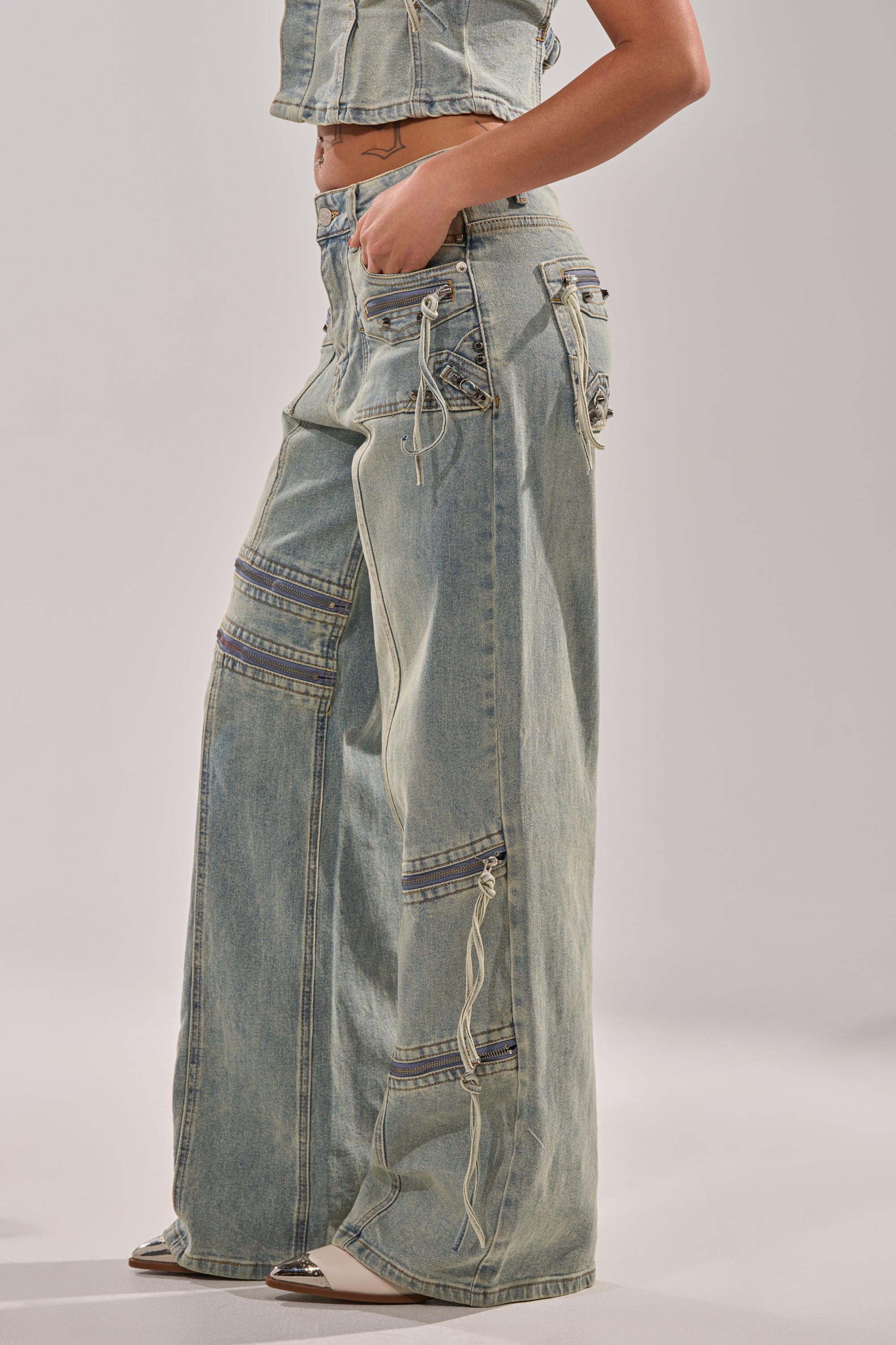 A person wears COMEBACK ERA WIDE LEG DENIM JEANS with decorative zippers, utility pockets, and stitching details, paired with a matching cropped denim top and pointed-toe shoes against a neutral, minimal background. 3