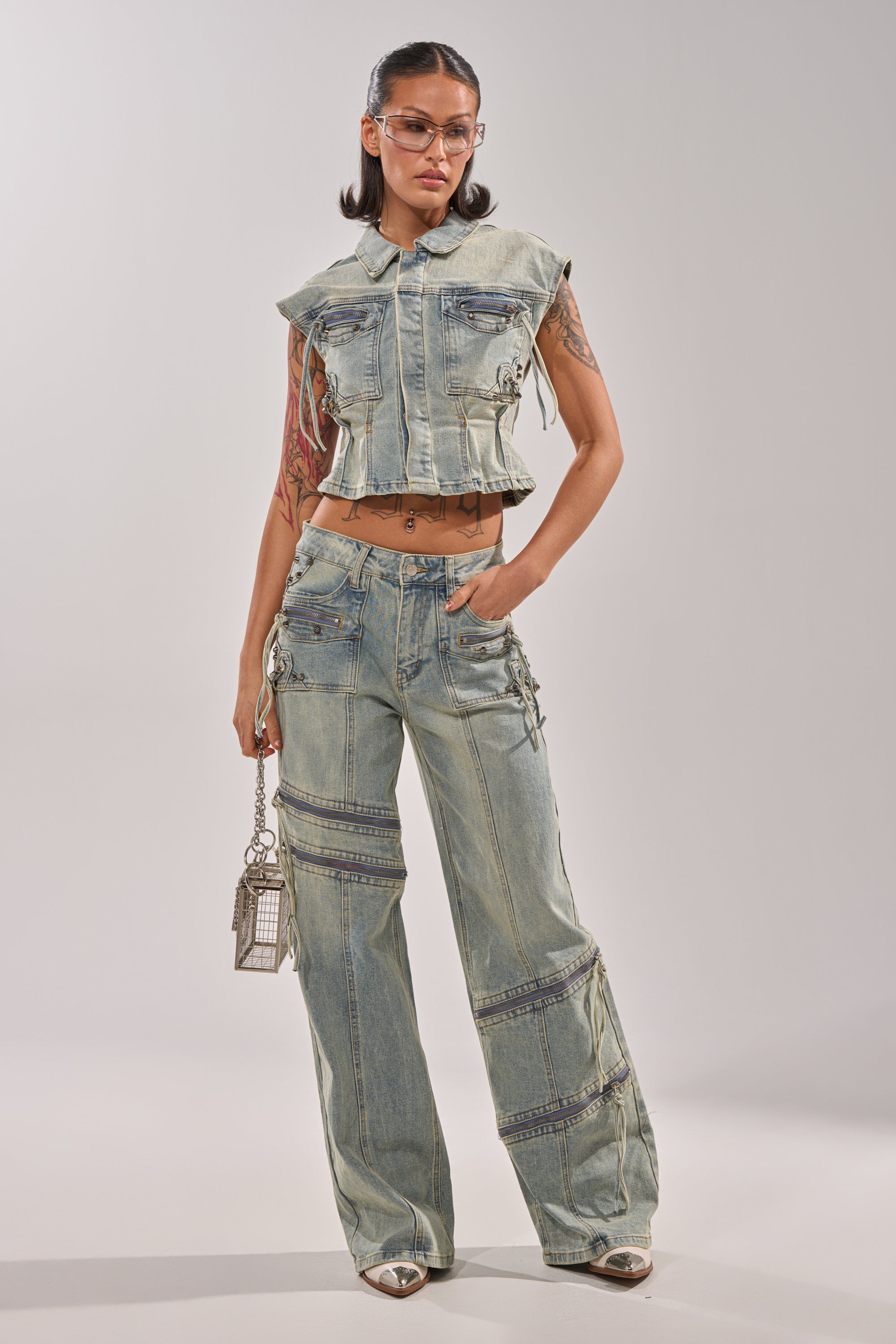 A woman stands confidently in COMEBACK ERA WIDE LEG DENIM JEANS and a sleeveless, cropped denim jacket with utility pockets. She wears glasses, silver pointed shoes, carries a small silver handbag, and has long dark hair and tattoos. 5