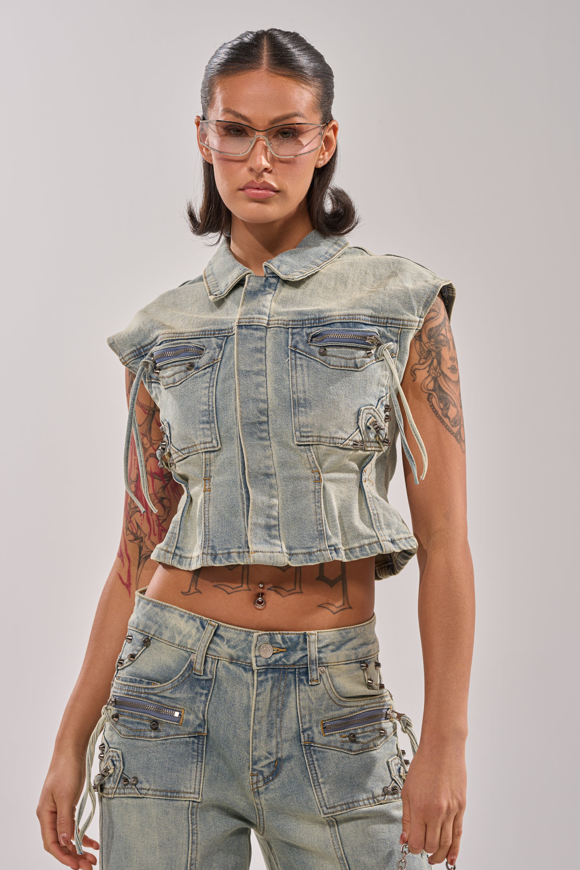 Wearing clear glasses and the COMEBACK ERA DENIM SLEEVELESS TOP, paired with matching jeans, a person showcases street style against a plain background, revealing tattoos on their arms and midriff. 0