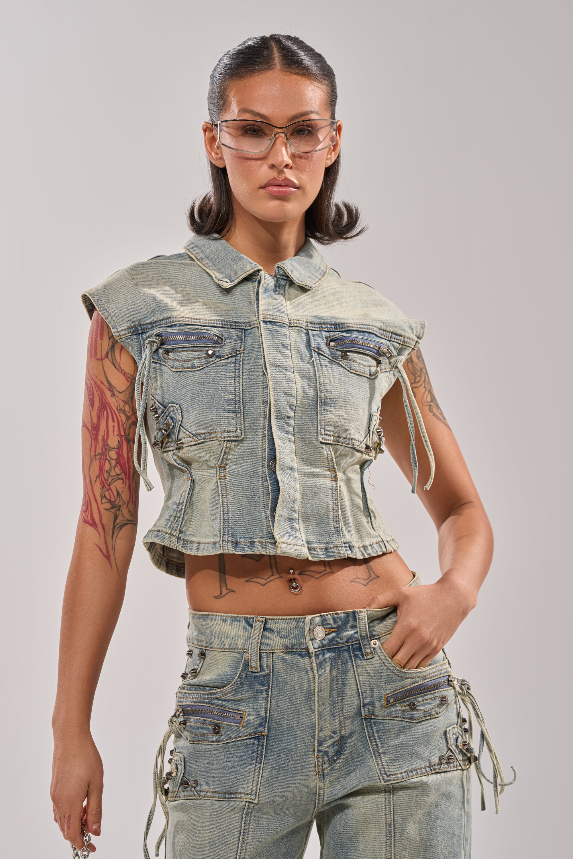 A woman with tattoos stands confidently with hand on hip, wearing the COMEBACK ERA DENIM SLEEVELESS TOP and matching jeans. Her slicked-back hair, clear glasses, and neutral expression complete a modern street style look against a plain background. 1